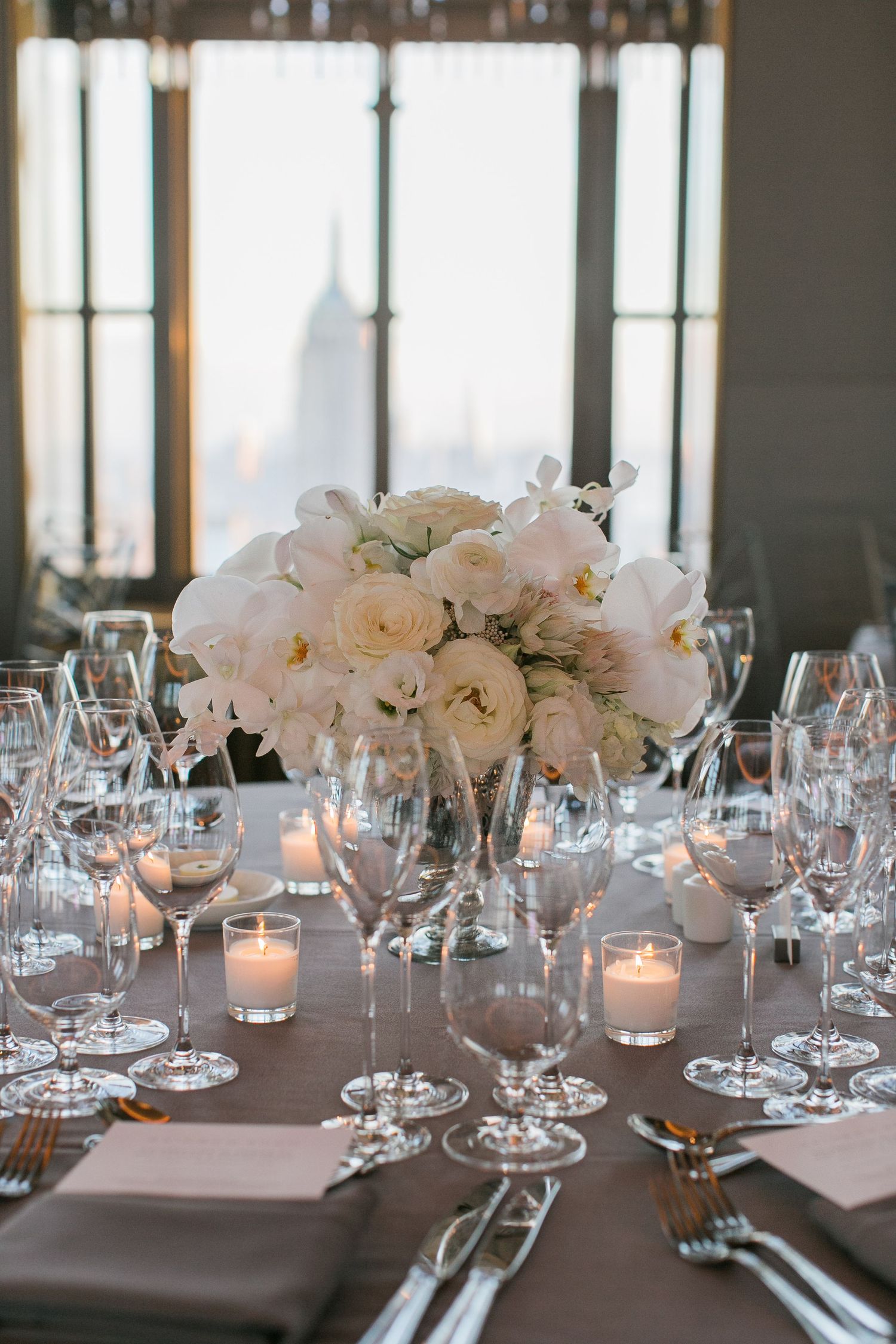 Elegant wedding reception table with white floral centerpiece, candles and wine glasses on gray tablecloth.