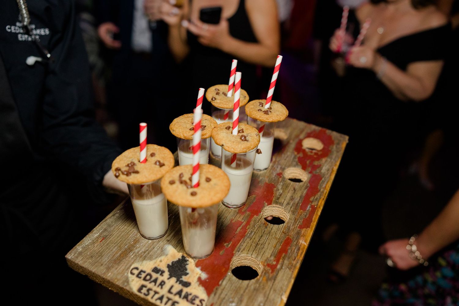 Wedding dessert display featuring mini milkshakes with cookies on a wooden serving board with candles.