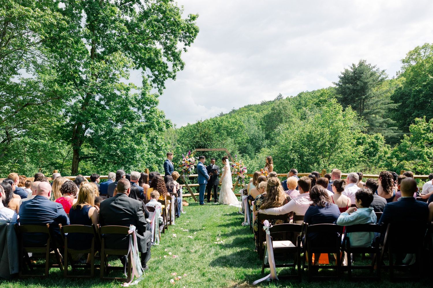 Outdoor wedding ceremony setup with rows of chairs on a grassy hillside surrounded by lush forest.