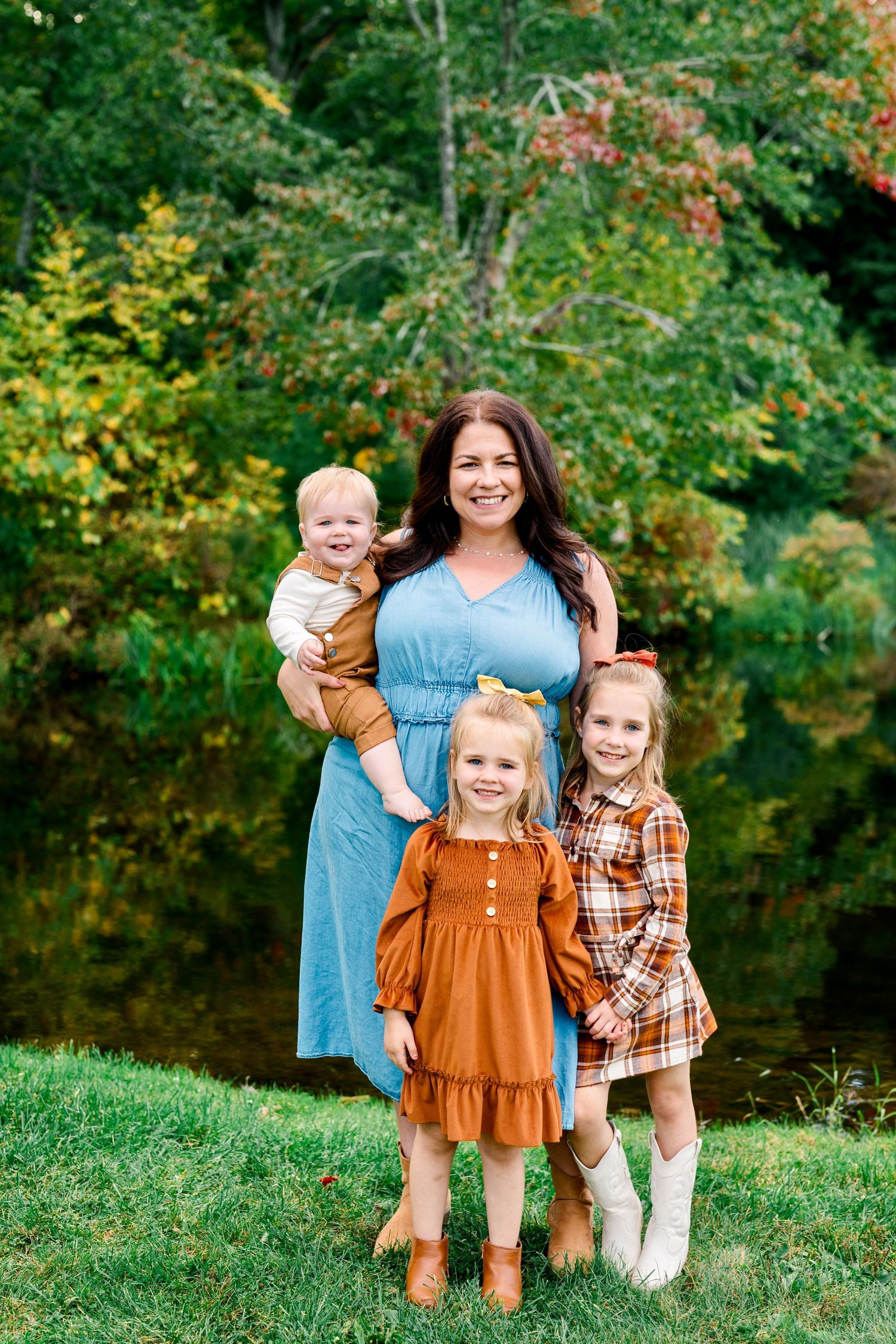 A smiling mother in a blue dress holds a baby while two young girls in fall outfits pose outdoors near a pond.