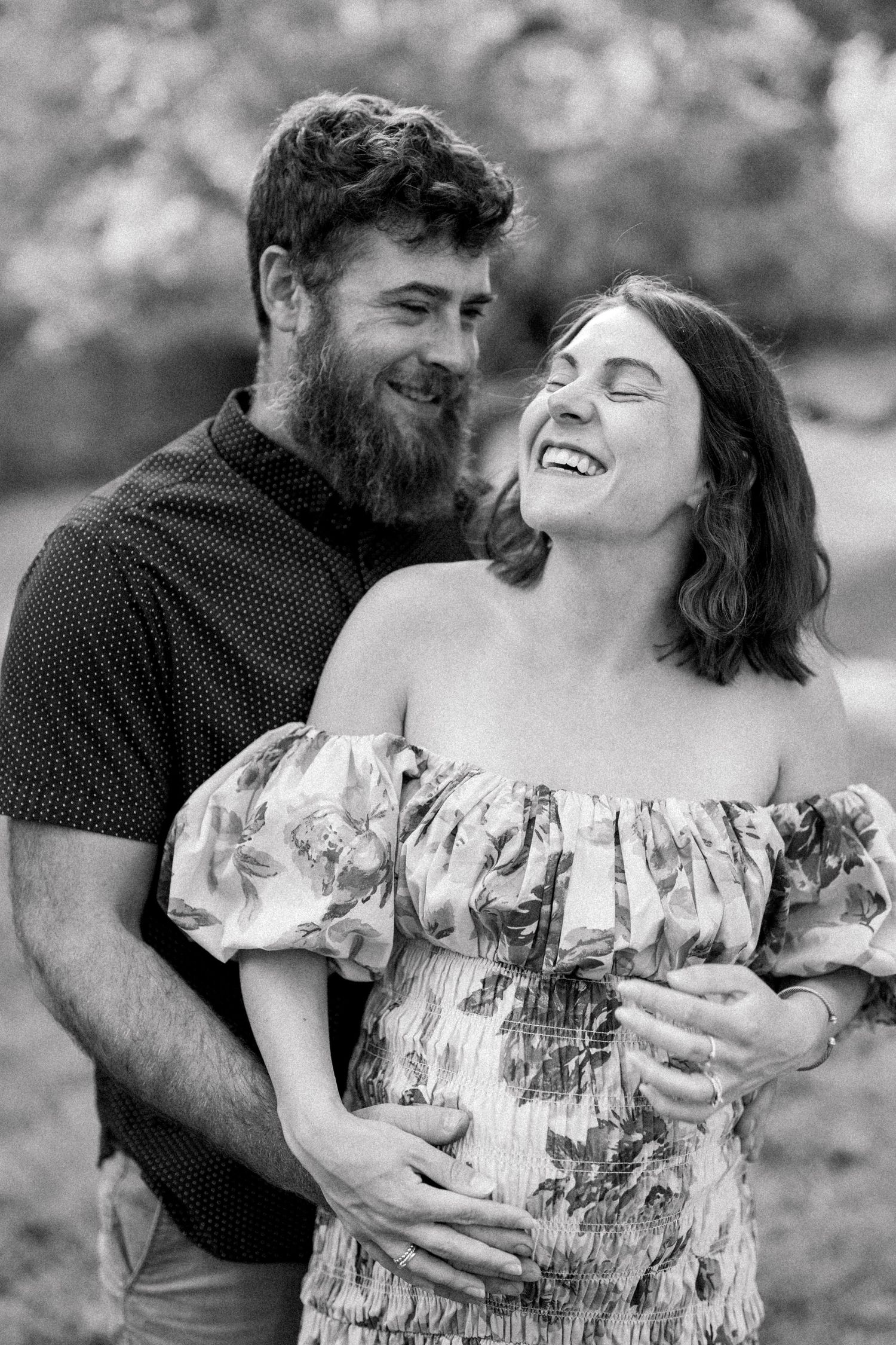 Black and white photo of a bearded man embracing a laughing pregnant woman outdoors.