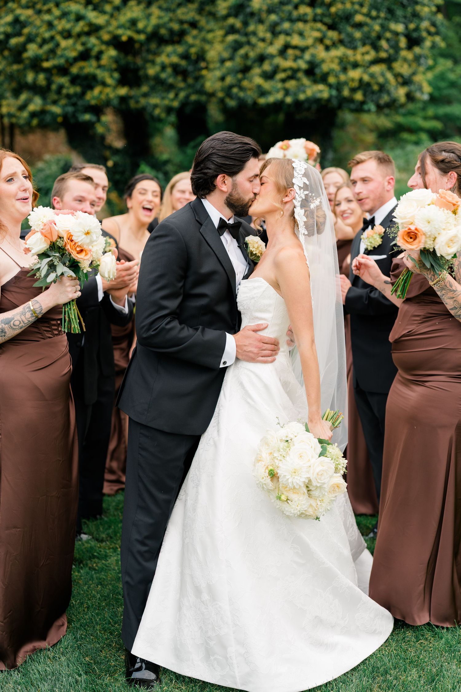 Bride and groom share a kiss surrounded by cheering bridesmaids in brown gowns and groomsmen at an outdoor wedding.