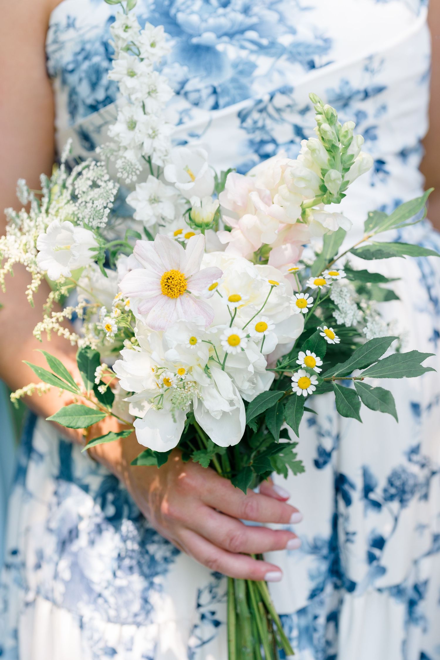 Woman in blue floral dress holding a white wildflower bouquet with daisies and cosmos blooms.