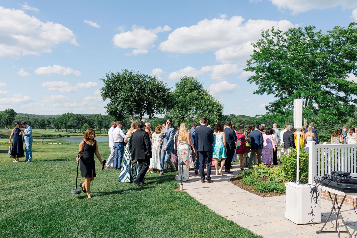 Wedding guests stroll along a garden pathway toward a scenic outdoor venue on a bright sunny day.