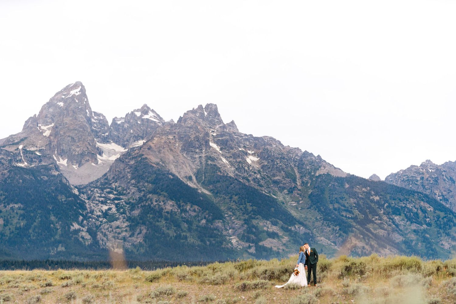 Wedding couple posing in a field with majestic Teton mountain peaks rising in the dramatic sunset landscape.