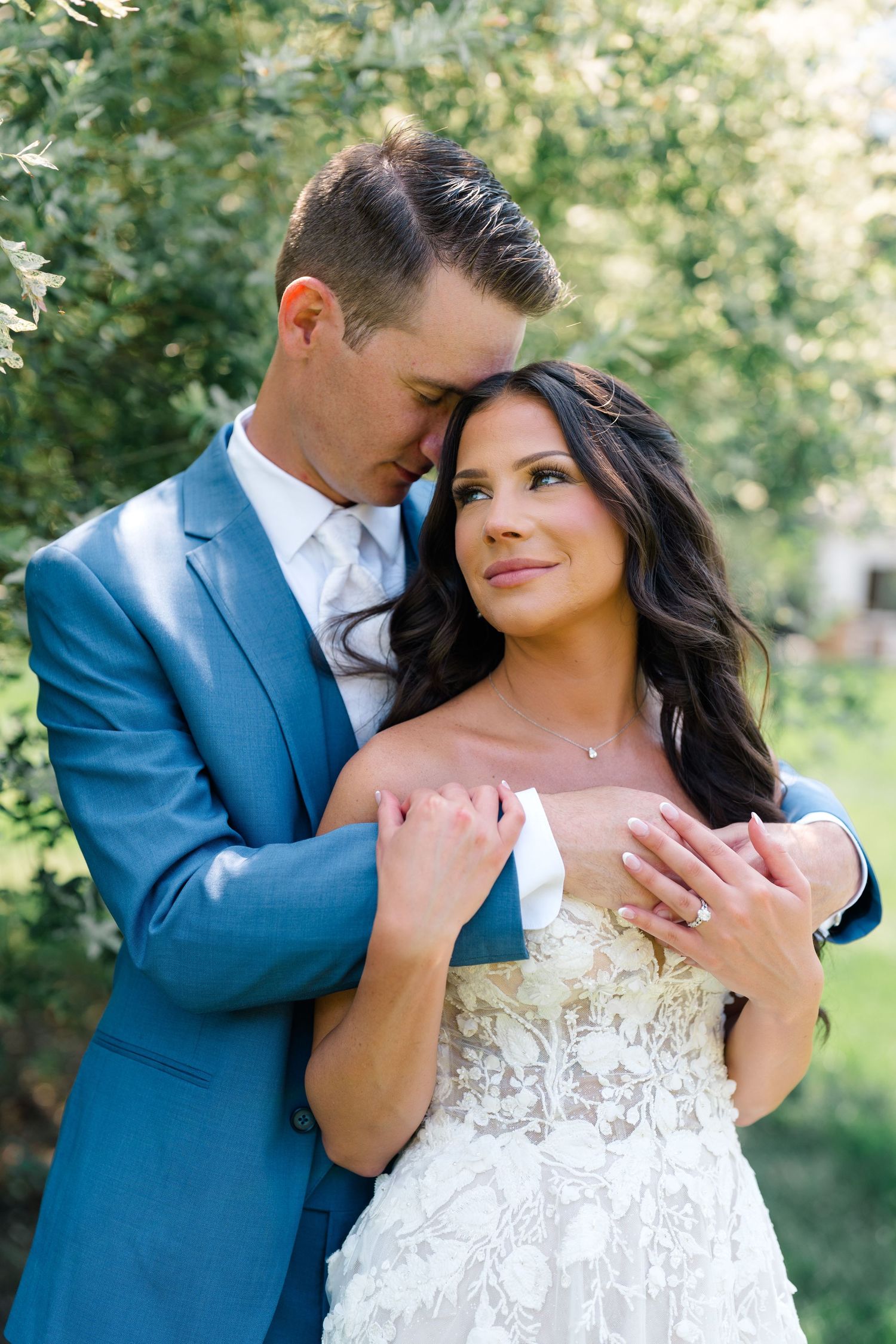 Bride in lace gown and groom in blue suit embrace during outdoor portrait session.