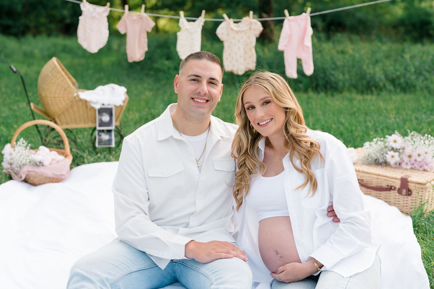 Maternity photoshoot in a garden setting with baby clothes on clothesline and picnic basket accessories.