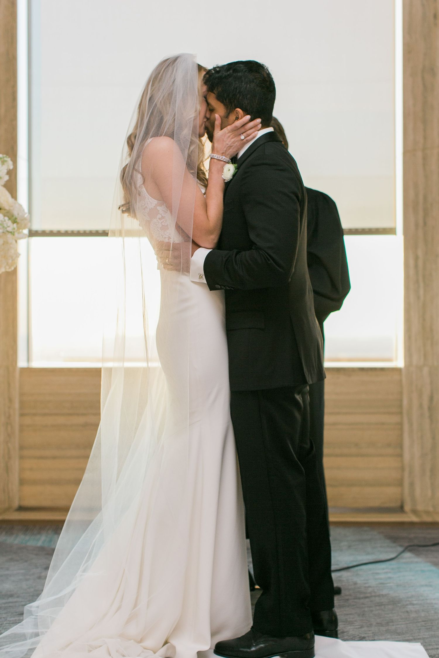Bride in sleek white dress and groom in classic black tuxedo share an intimate kiss by a sunlit window.