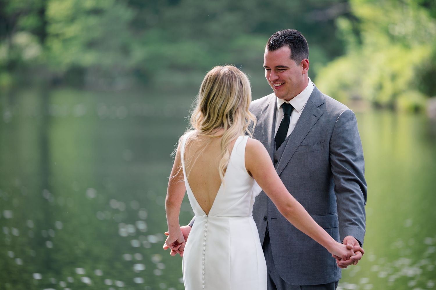 A couple shares a romantic moment by a tranquil lake with lush greenery, bride in white gown and groom in gray suit.