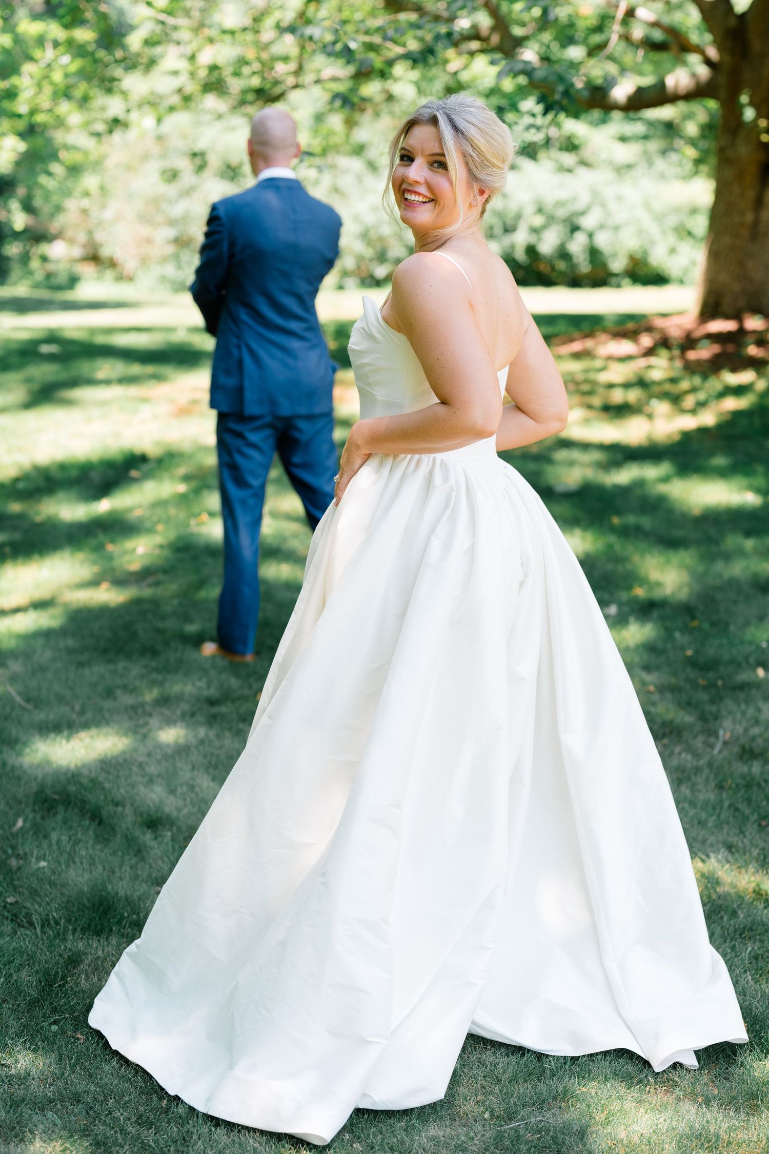 Smiling bride in strapless white ball gown looks over shoulder outdoors while groom in navy suit stands ahead.