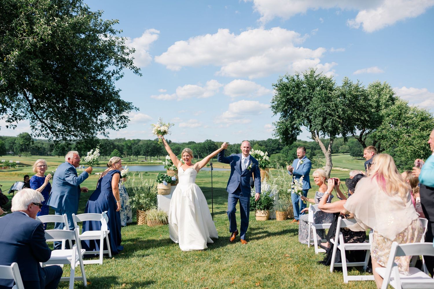 Jubilant newlyweds raise their arms in celebration while exiting their outdoor ceremony past cheering guests.