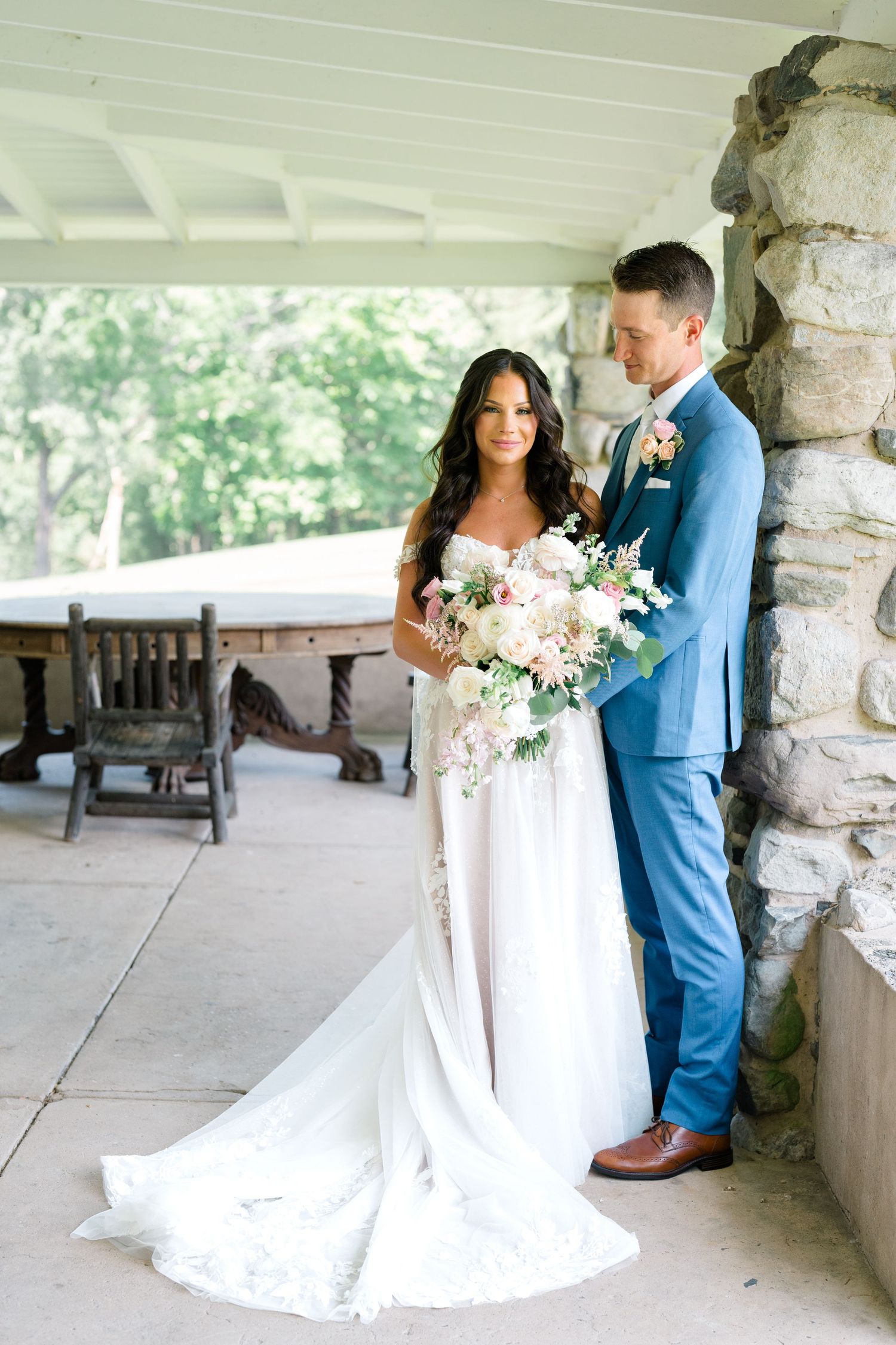 Bride in white gown and groom in blue suit embrace under stone archway with floral bouquet.