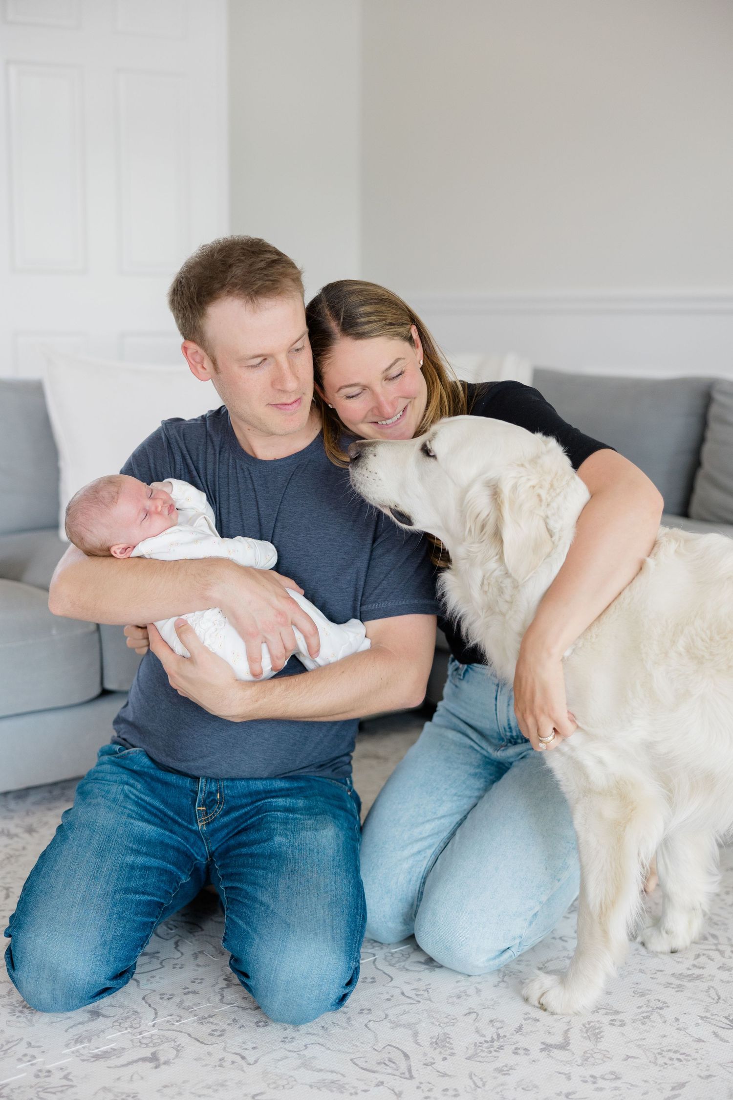 Candid family moment on floor with white dog and newborn baby in light-filled room.