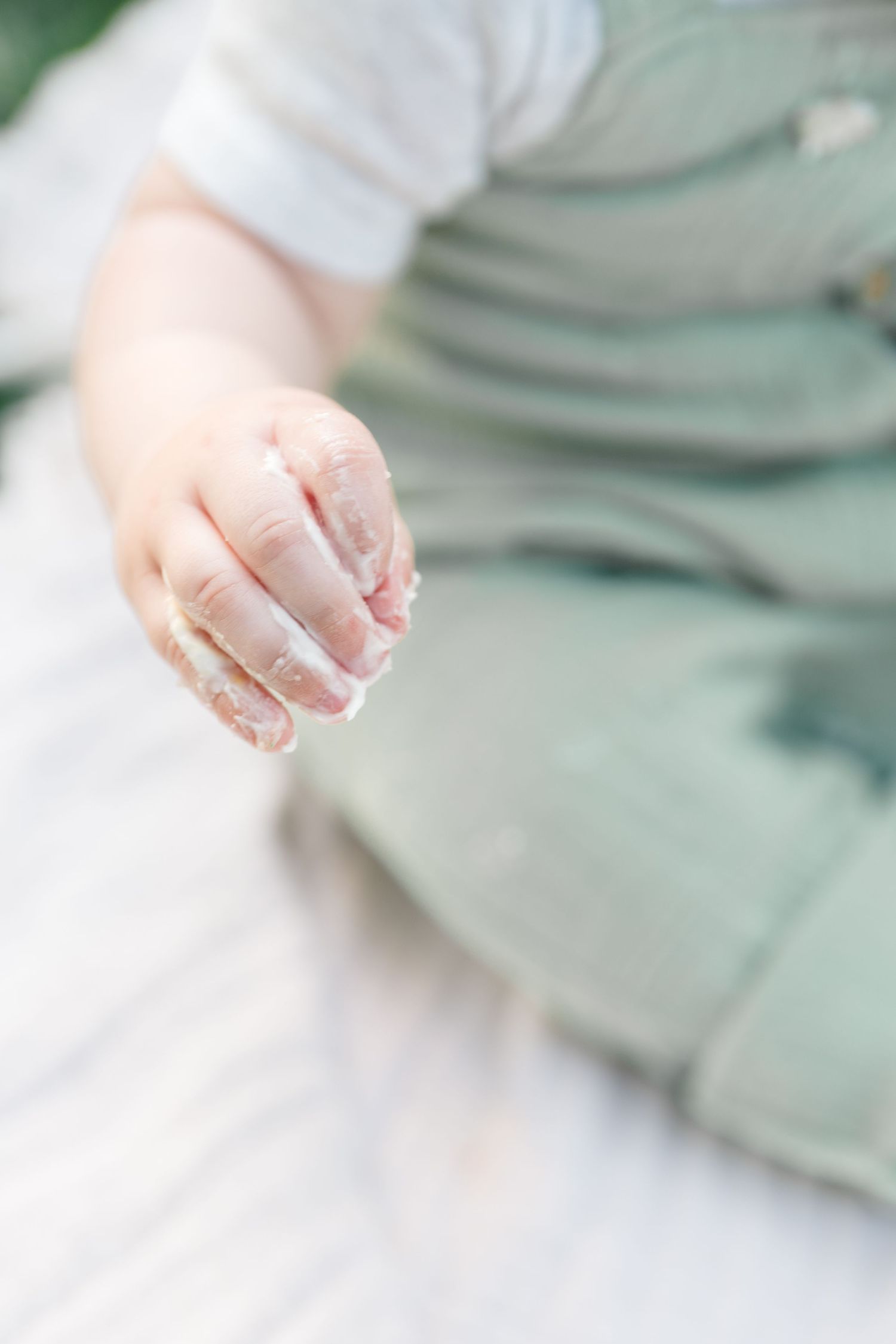 Close-up of a baby's hand covered in white cake frosting, wearing sage green overalls during a first birthday smash cake.