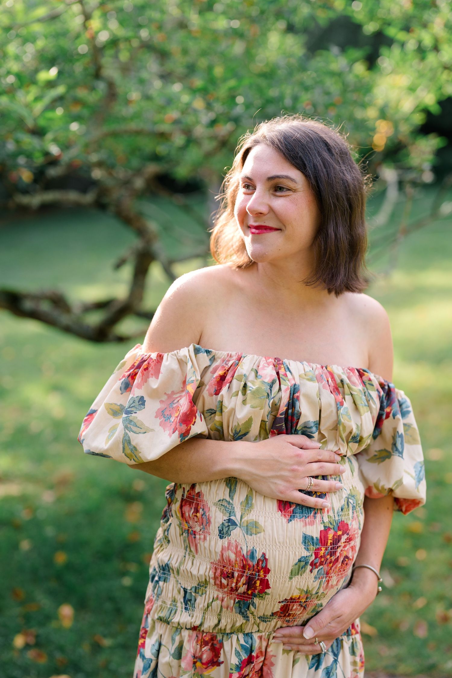 Pregnant woman smiling in floral off-shoulder dress, cradling baby bump in lush green garden setting.