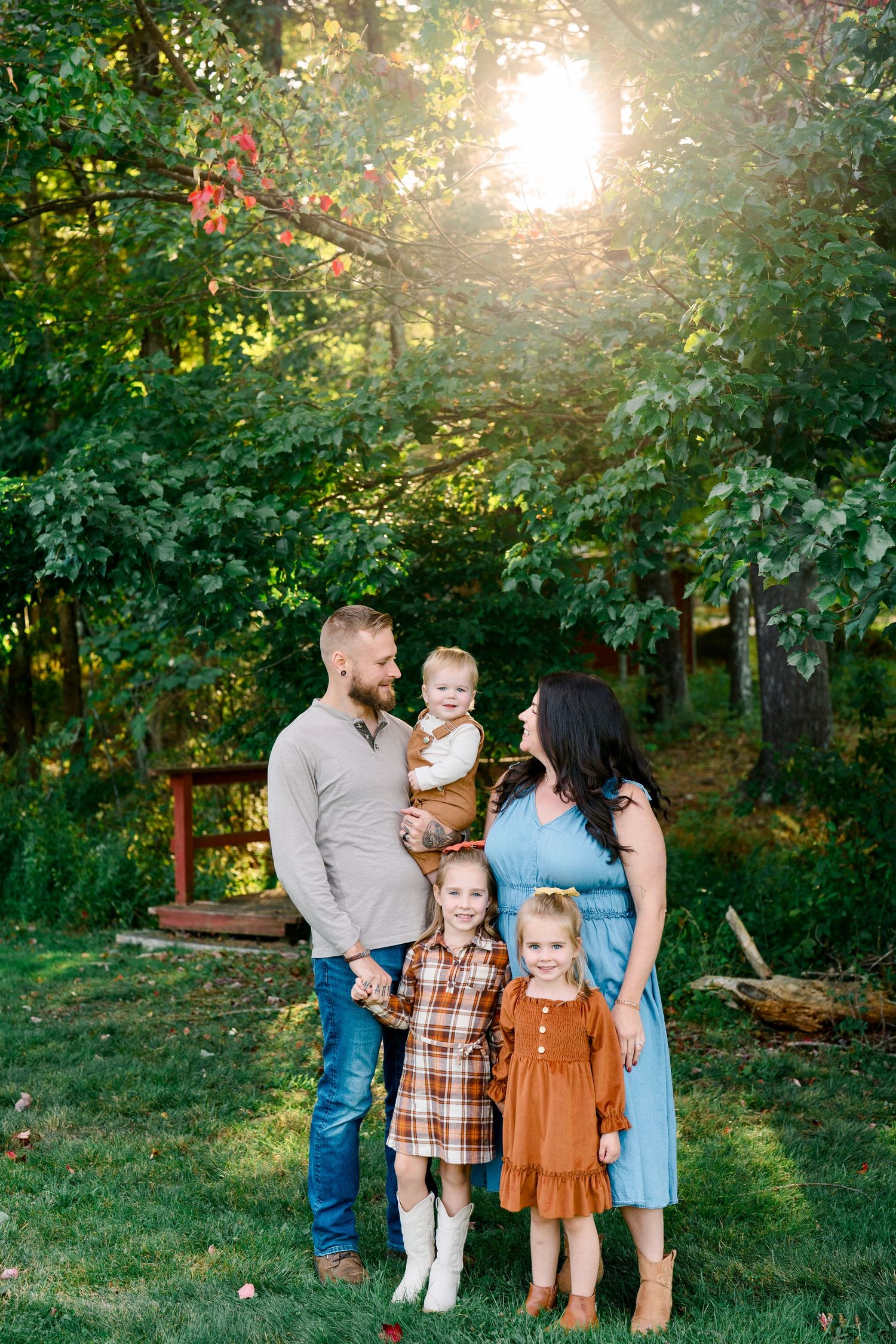 Family of five posing outdoors in fall outfits with golden sunlight filtering through lush green trees.