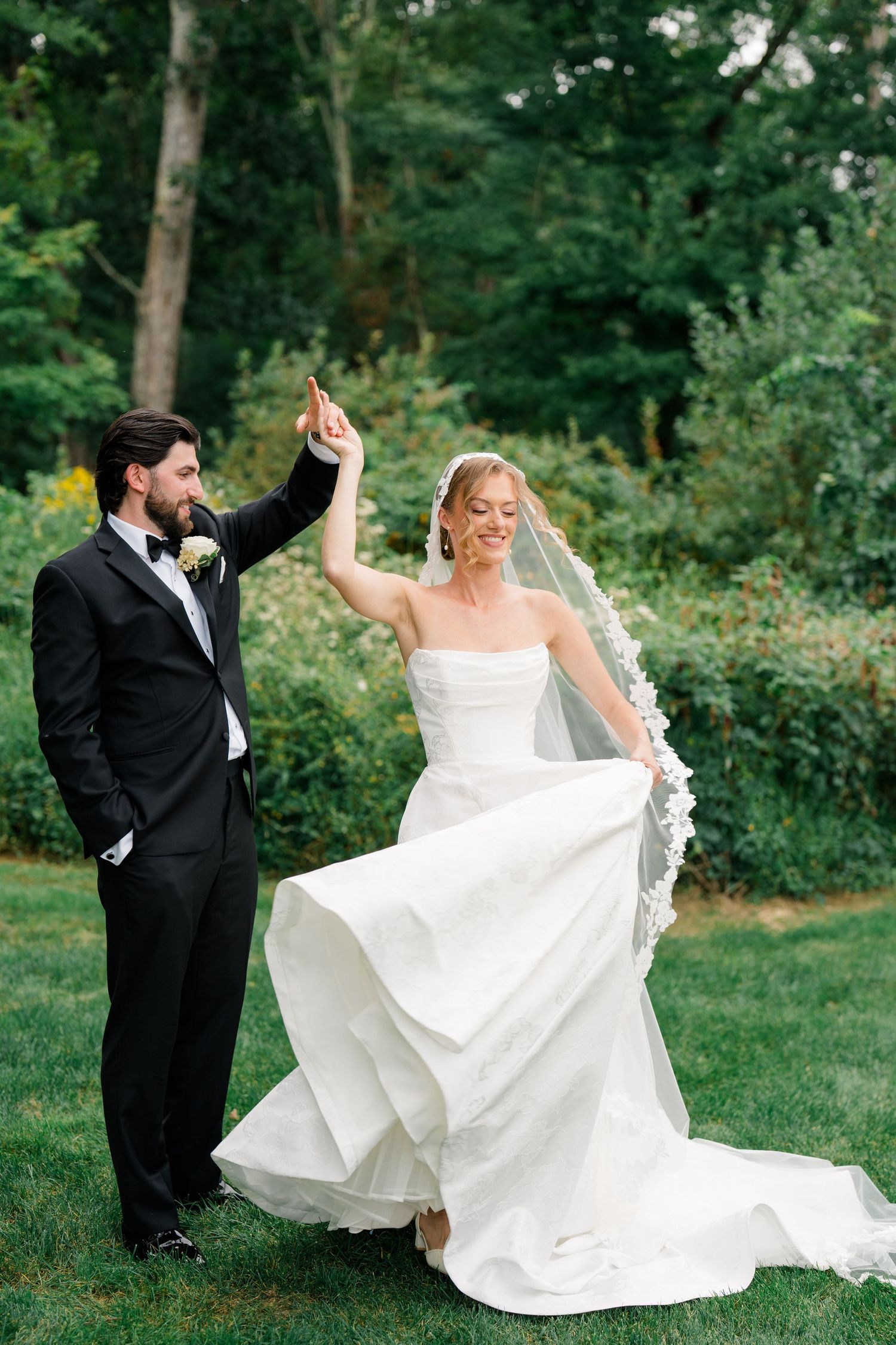 Groom twirls bride in flowing white strapless gown and lace veil on green lawn with garden backdrop.