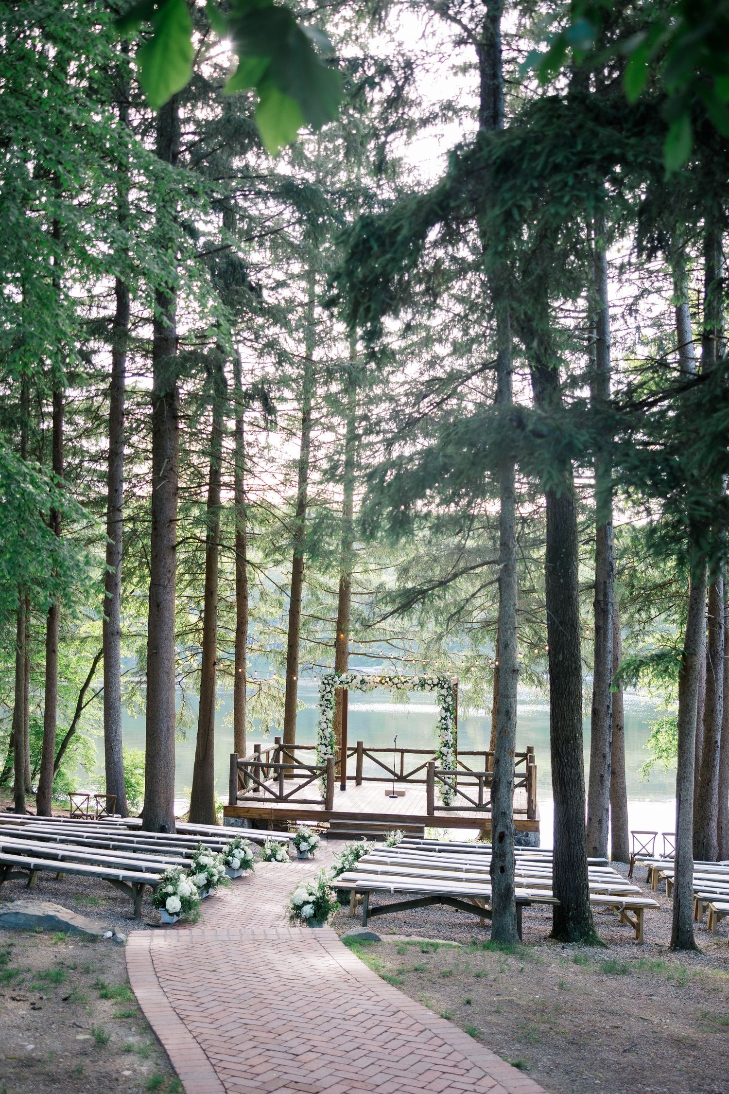 Brick pathway leads through pine forest to lakeside wooden gazebo with rustic bench seating for outdoor ceremonies.