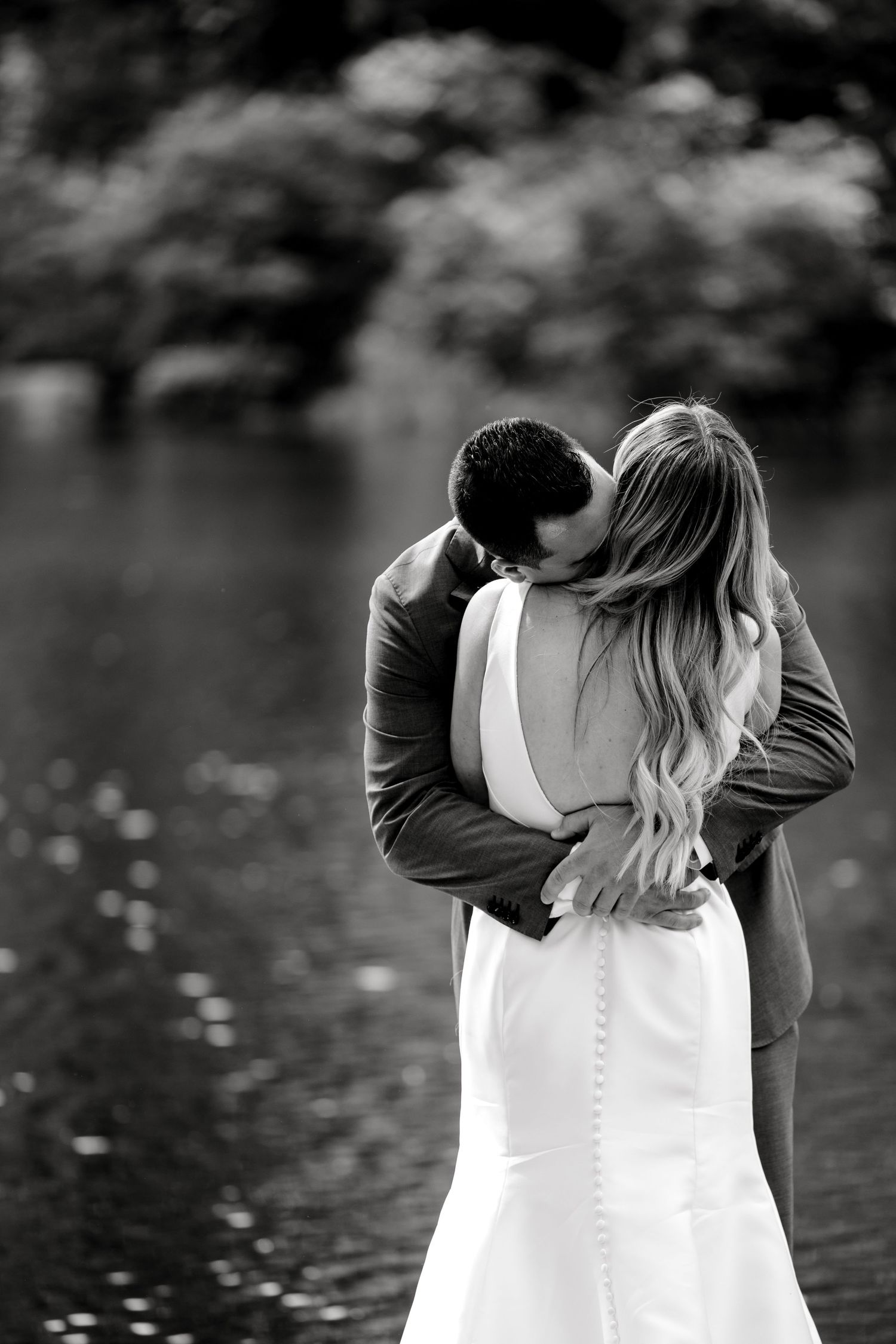 A romantic black and white photograph of a couple embracing by a lake, with the bride wearing a white wedding dress.