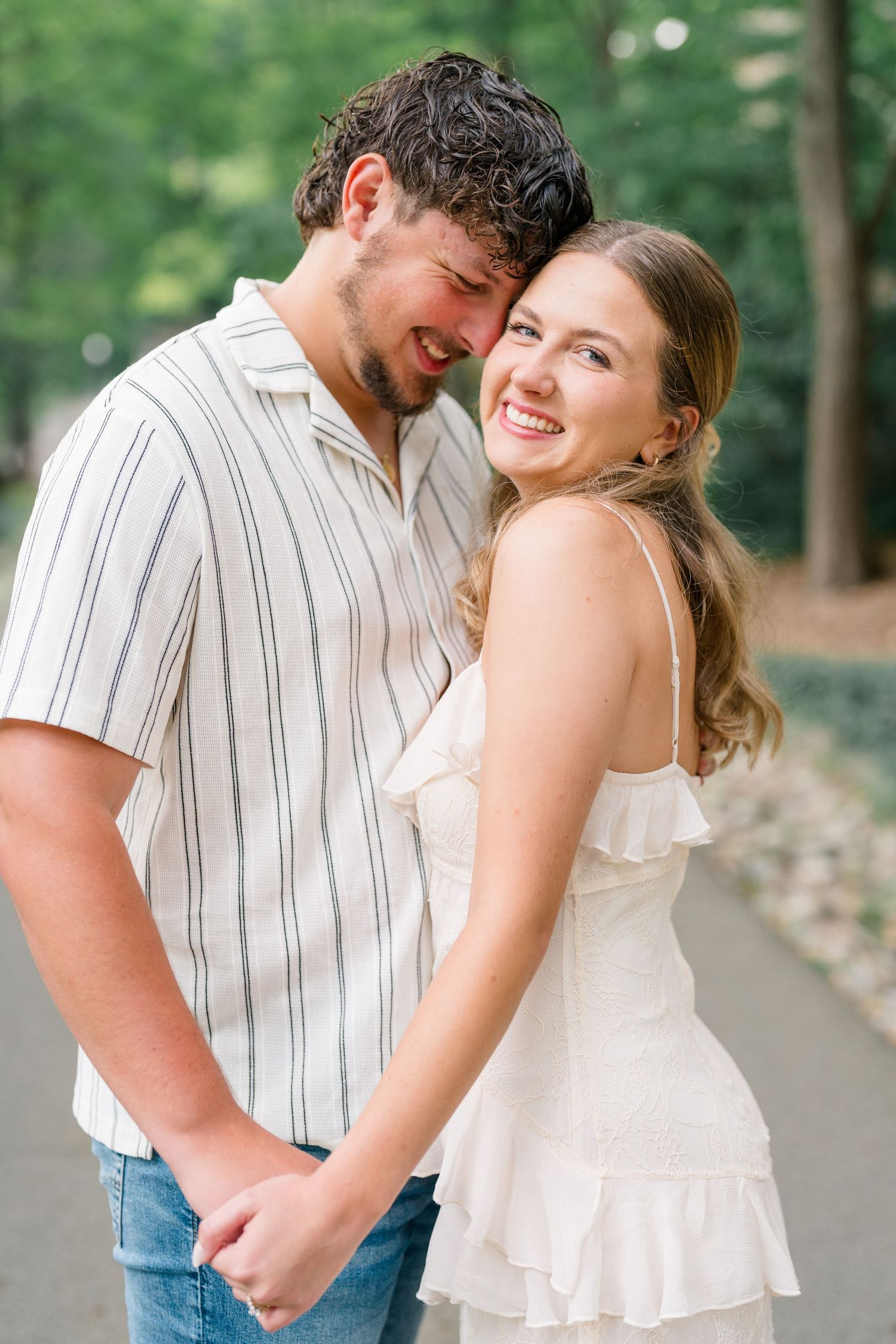 Smiling couple holding hands outdoors, woman in white ruffled dress laughing as man nuzzles her cheek on a tree-lined path.
