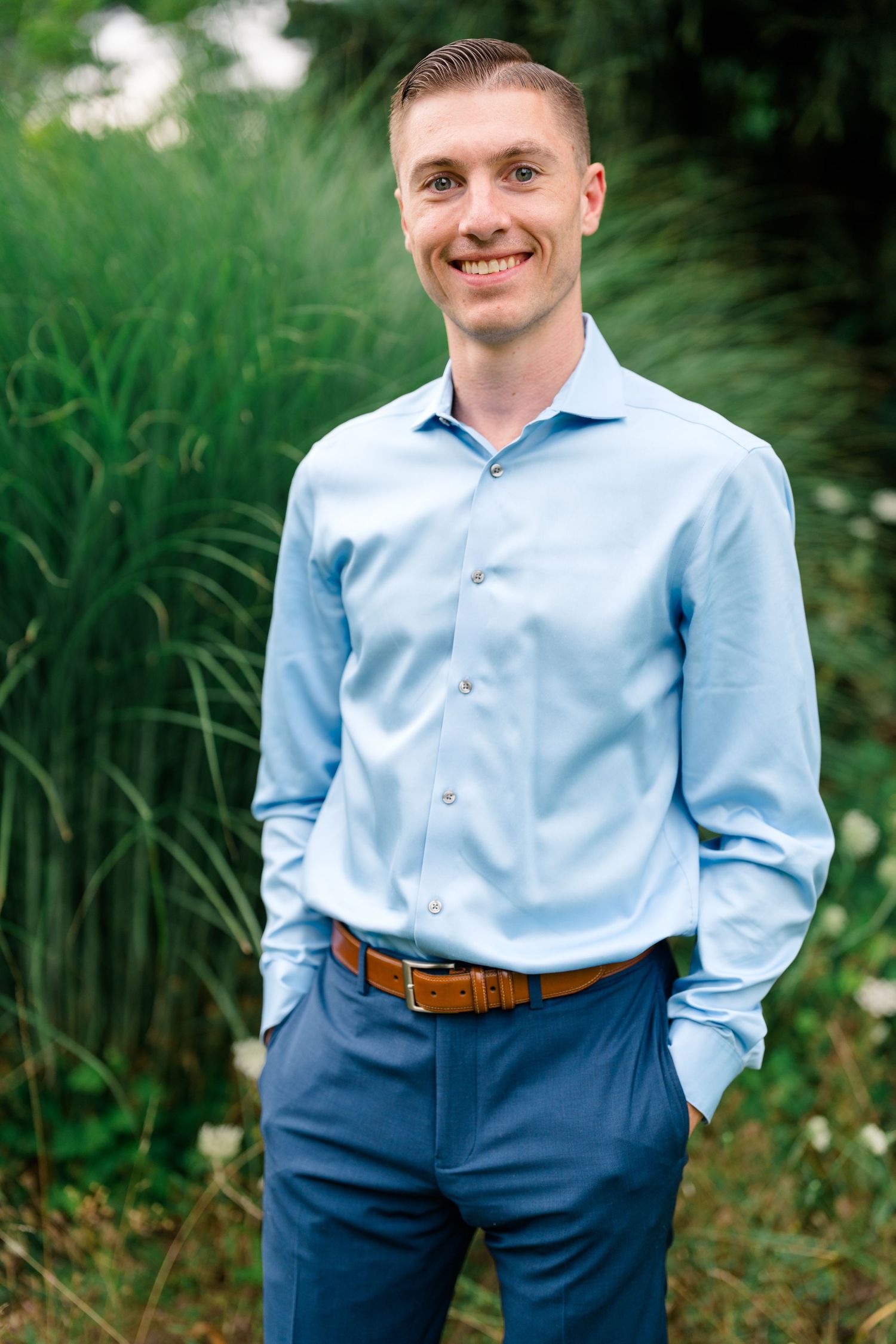 Smiling young man in blue dress shirt and navy pants standing outdoors in a garden setting.