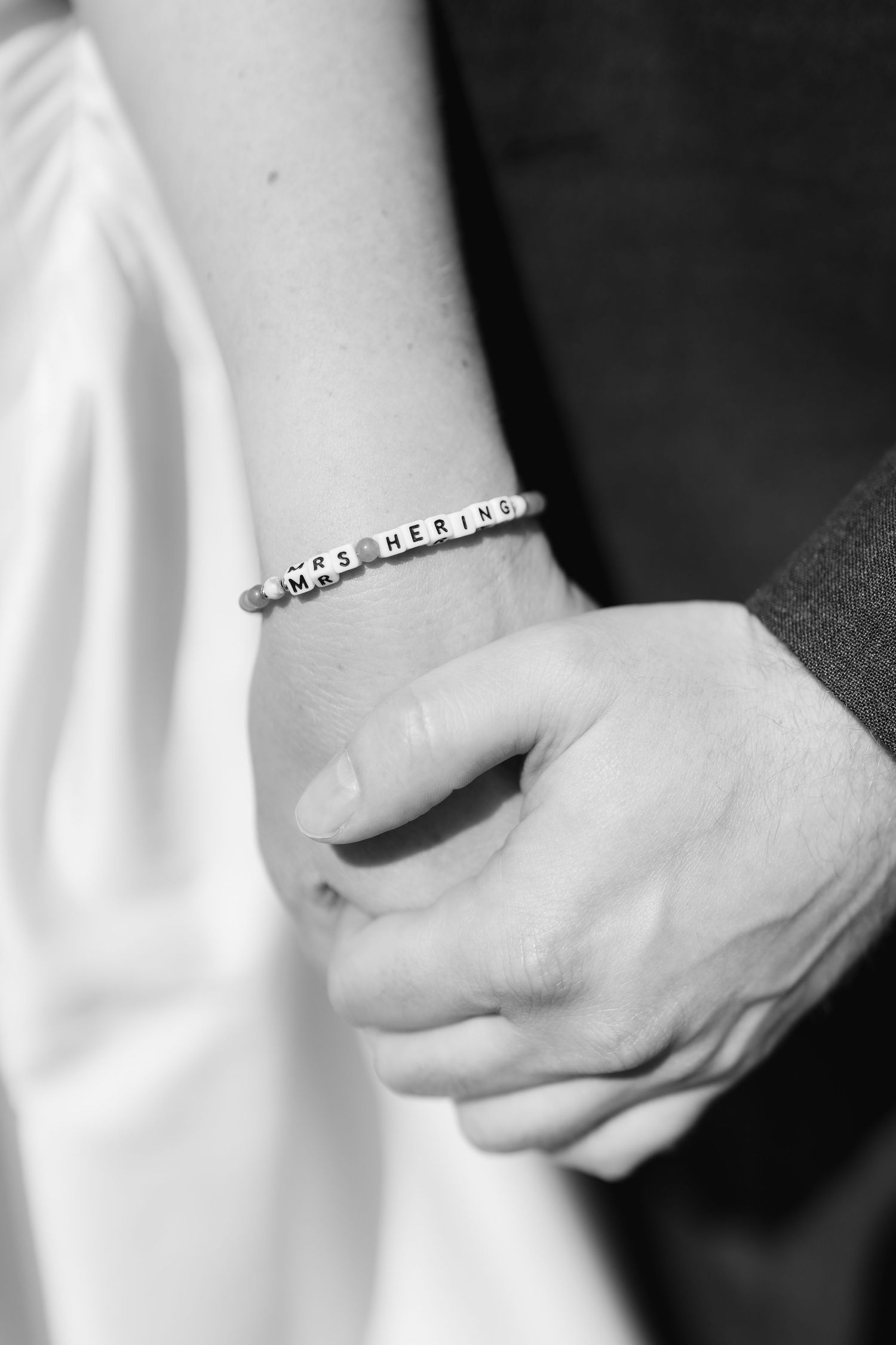Close-up of a bride's wrist wearing a letter bead bracelet reading MRS HERING while holding hands with groom.