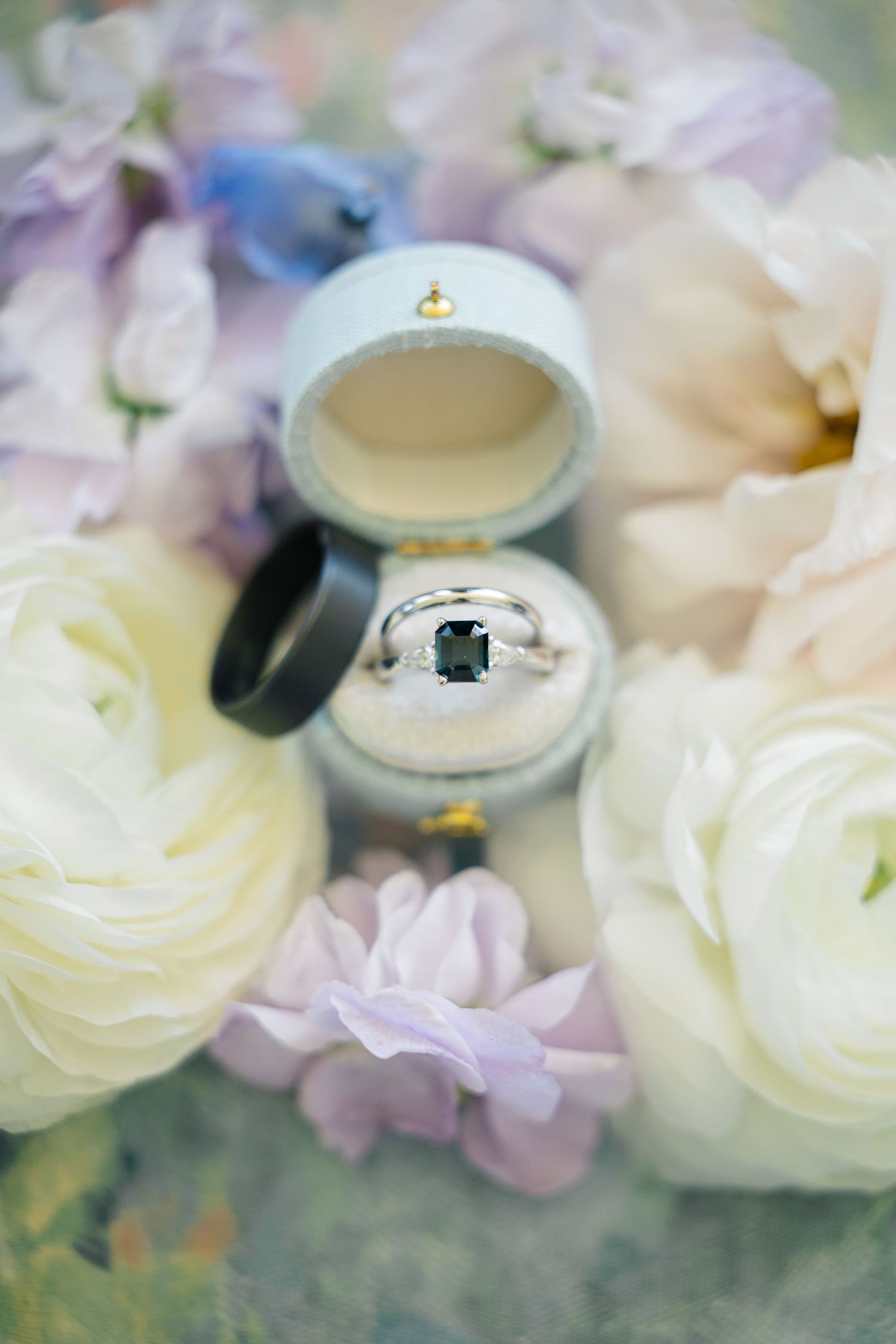 Delicate floral arrangement with white ranunculus flowers and soft pastel blooms surrounds vintage-style ring box in dreamy lighting.