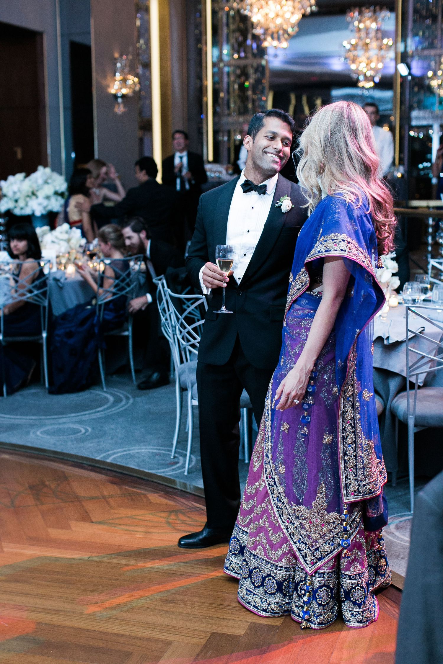 A couple at an elegant reception with the woman wearing an ornate purple gown while holding drinks in a chandelier-lit room.