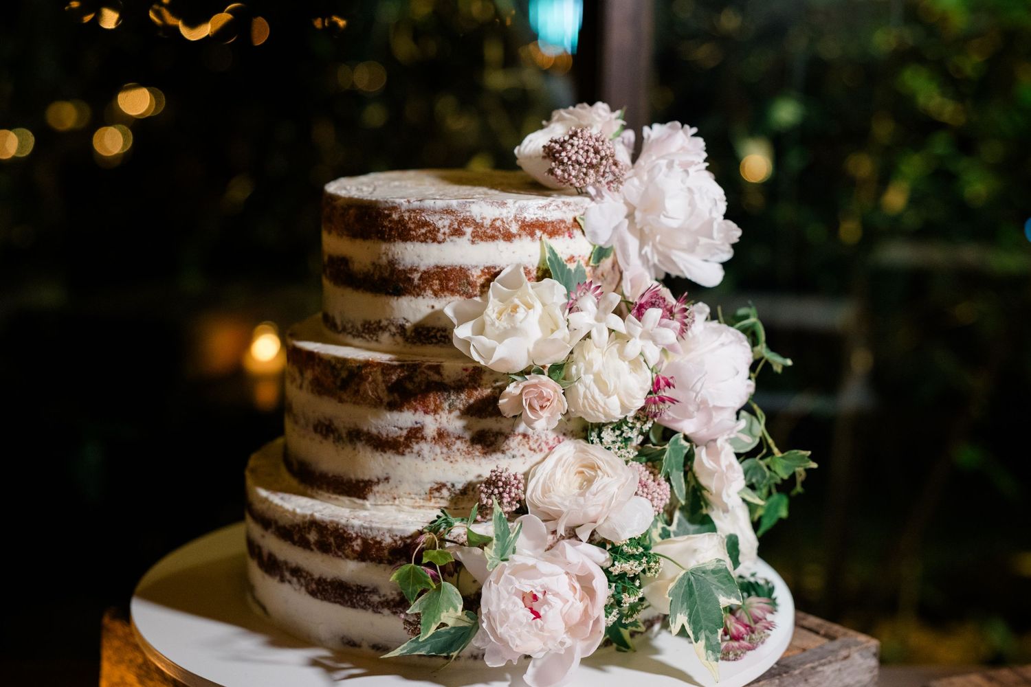 A rustic three-tier naked wedding cake decorated with fresh pink peonies and greenery on a white stand.