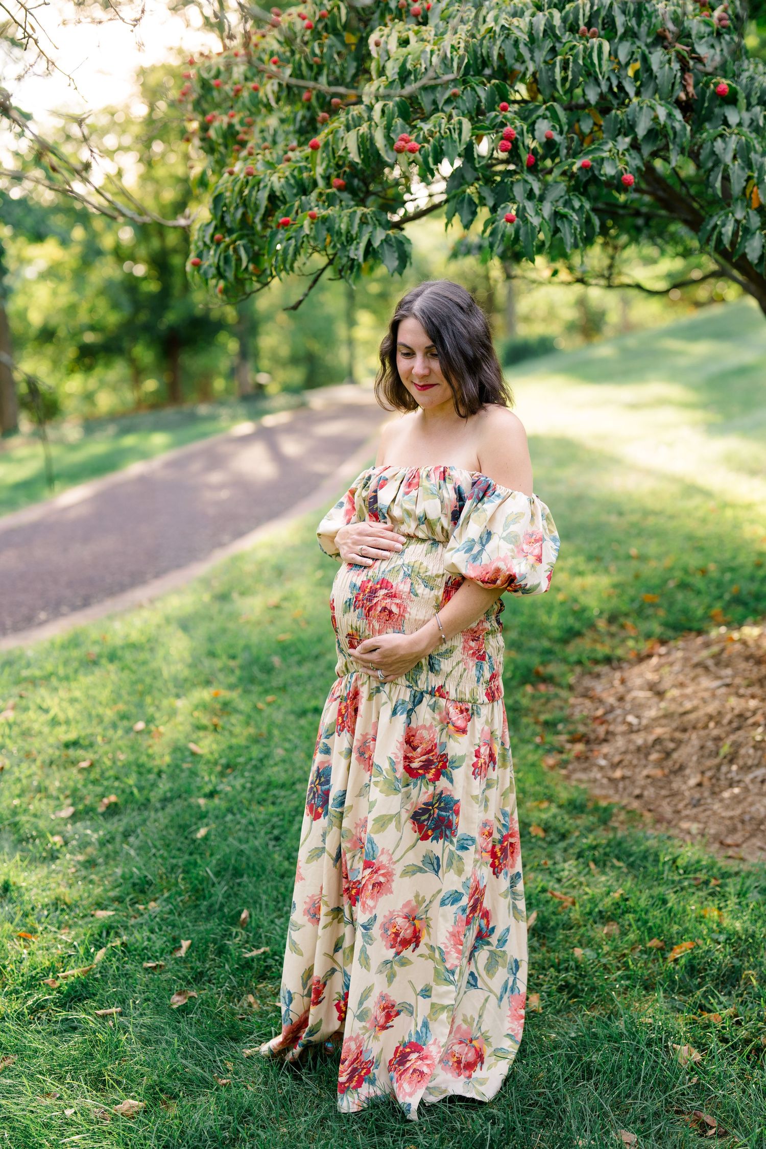 Pregnant woman in floral off-shoulder maxi dress cradling baby bump outdoors in lush green park setting.