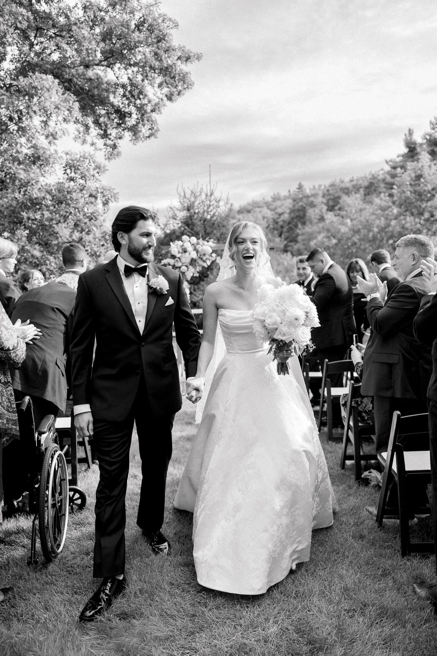 Black and white photo of a joyful bride and groom walking down the aisle at an outdoor wedding ceremony.