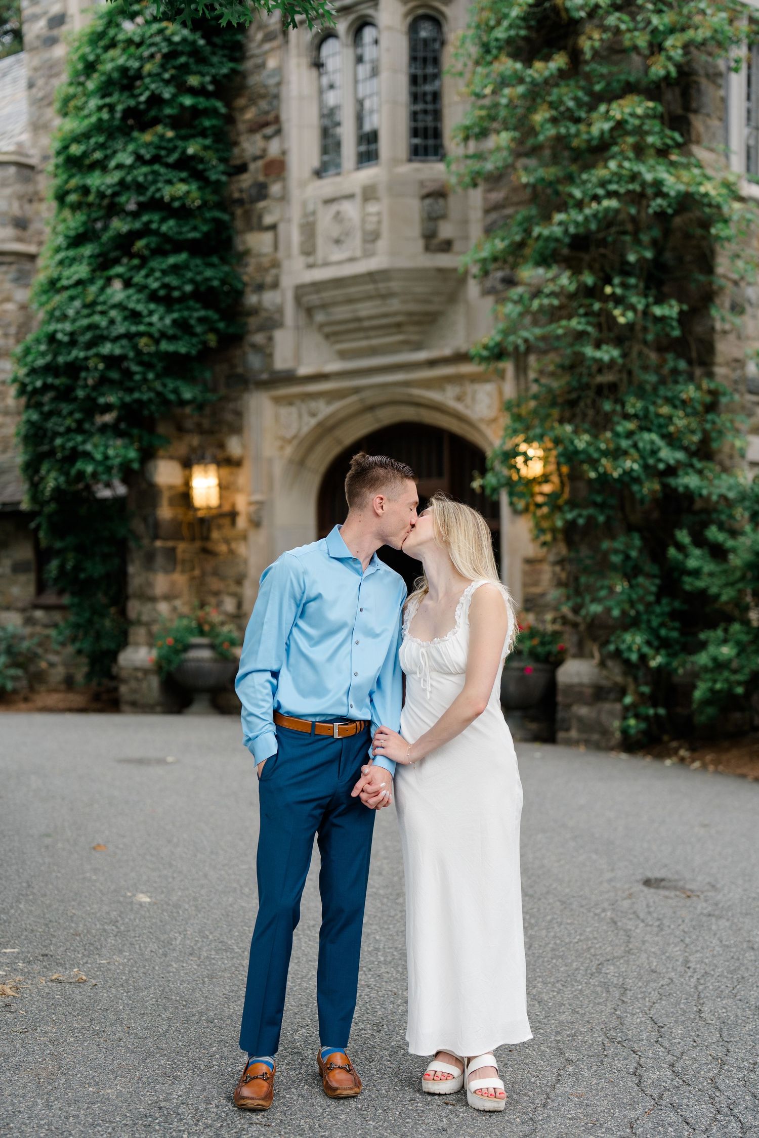 Couple sharing a kiss while holding hands in front of an ivy-covered stone building during their engagement session.