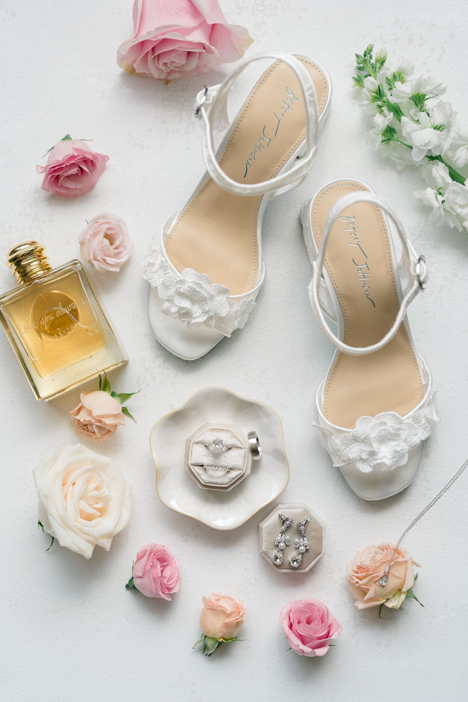Elegant bridal accessories including white shoes, perfume bottles, and delicate pink roses arranged on white surface.