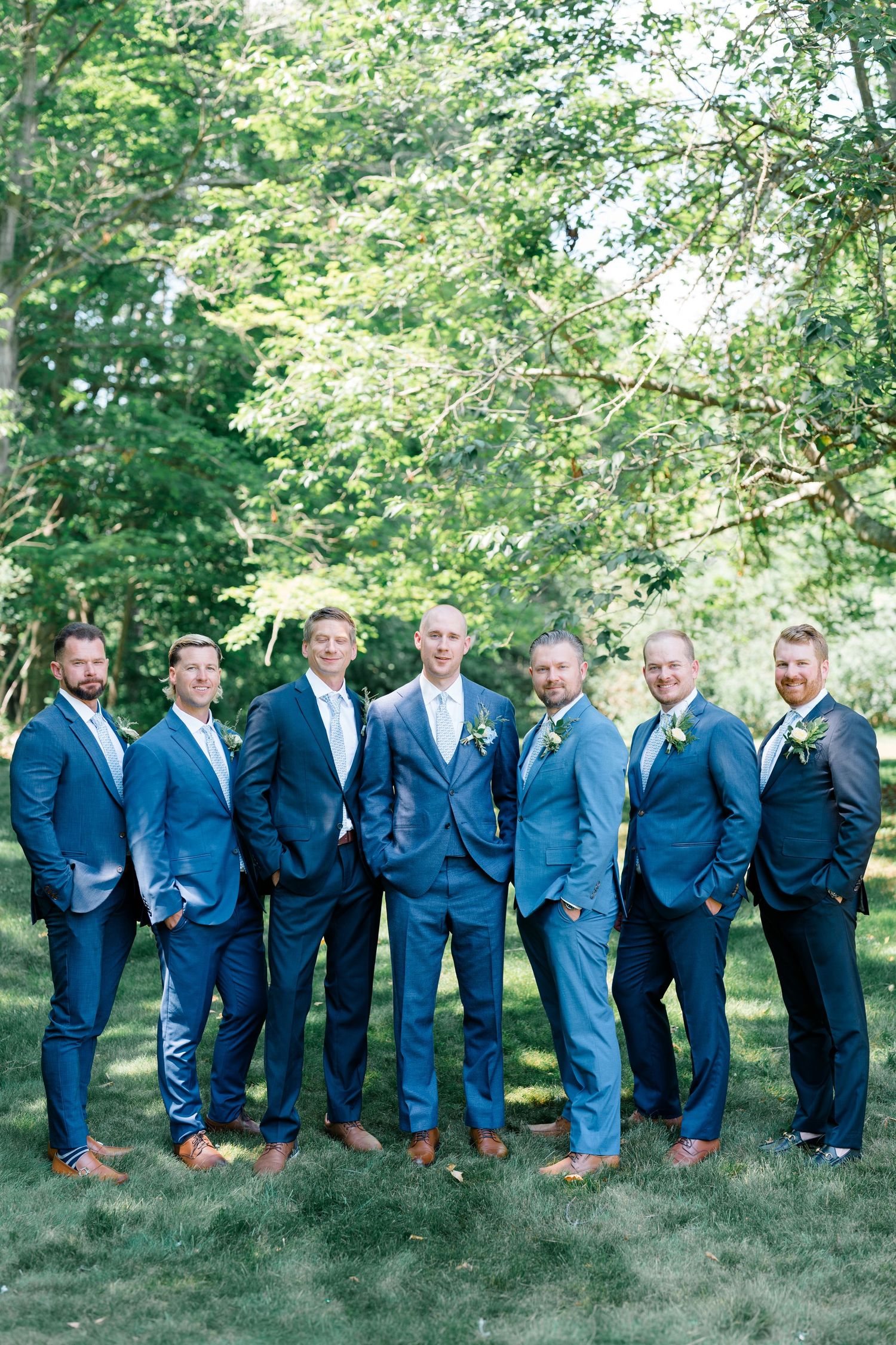 Seven groomsmen in matching blue suits pose together outdoors on green grass with lush trees in the background.