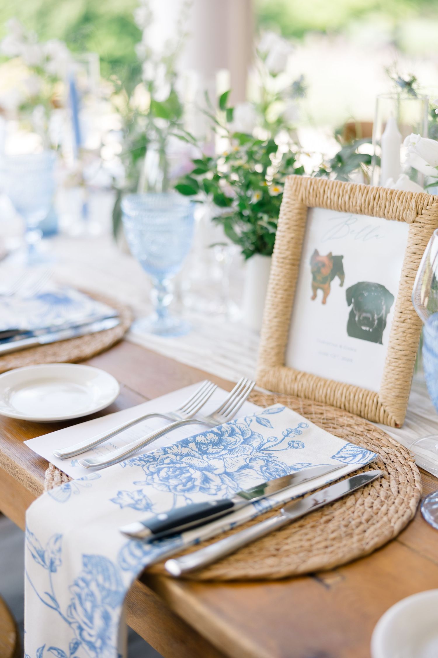 An elegant wedding table setting with blue floral napkins, wicker chargers, and a framed dog illustration.