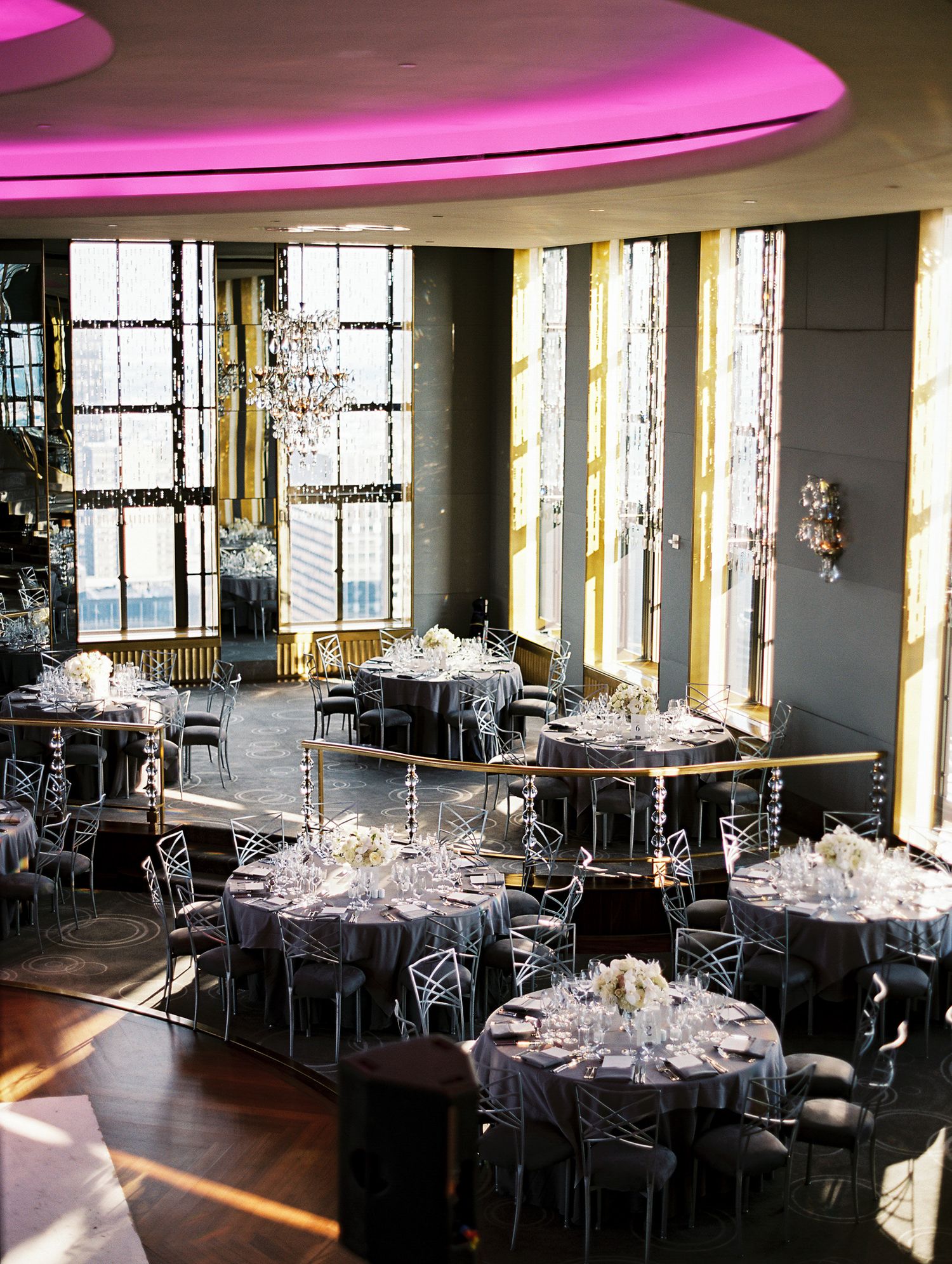Elegant wedding reception venue with round tables, crystal glassware, and dramatic floor-to-ceiling windows under pink mood lighting.