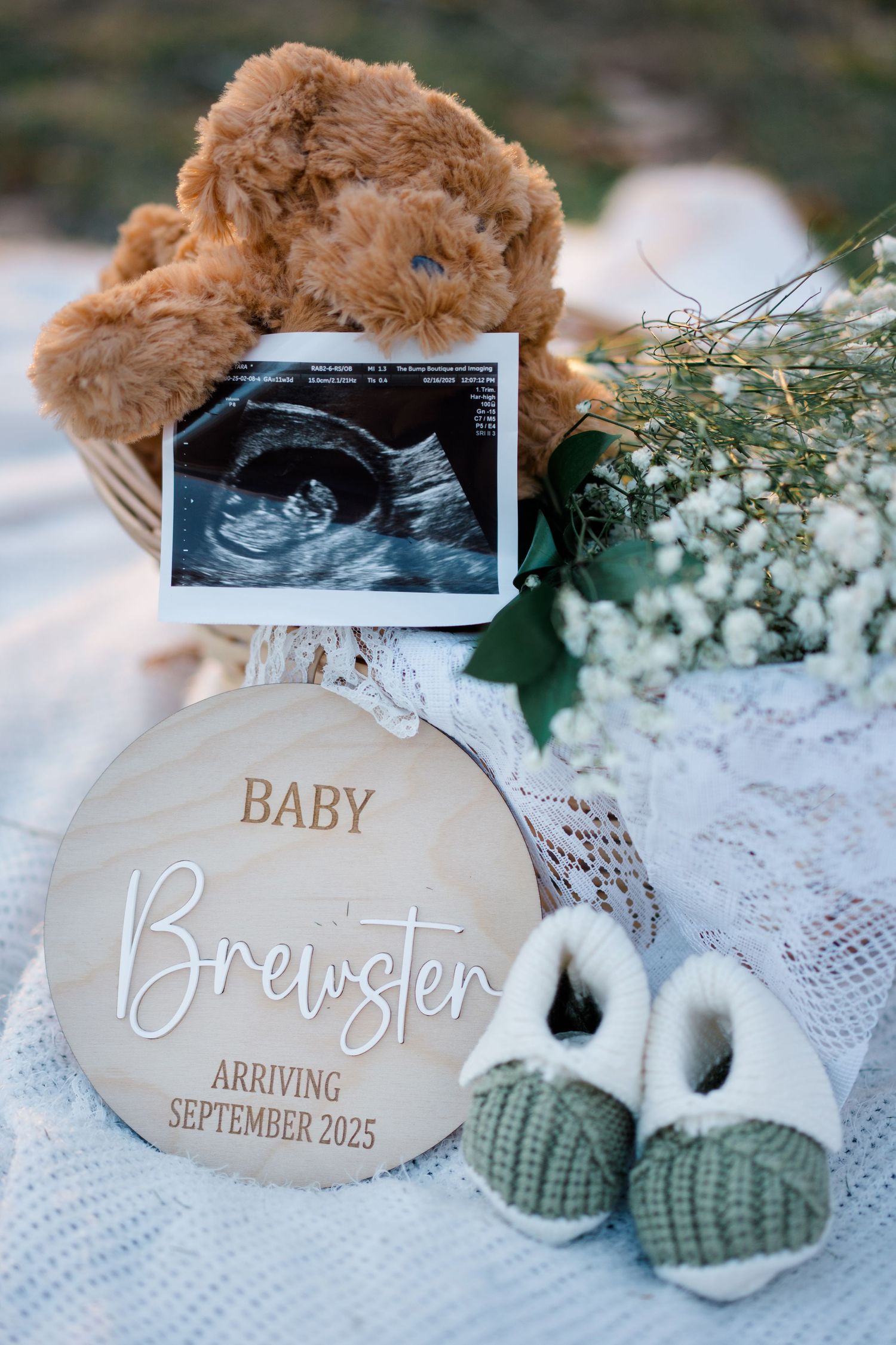 Pregnancy announcement featuring ultrasound photo, teddy bear, tiny booties, and wooden name sign for Baby Brewster.