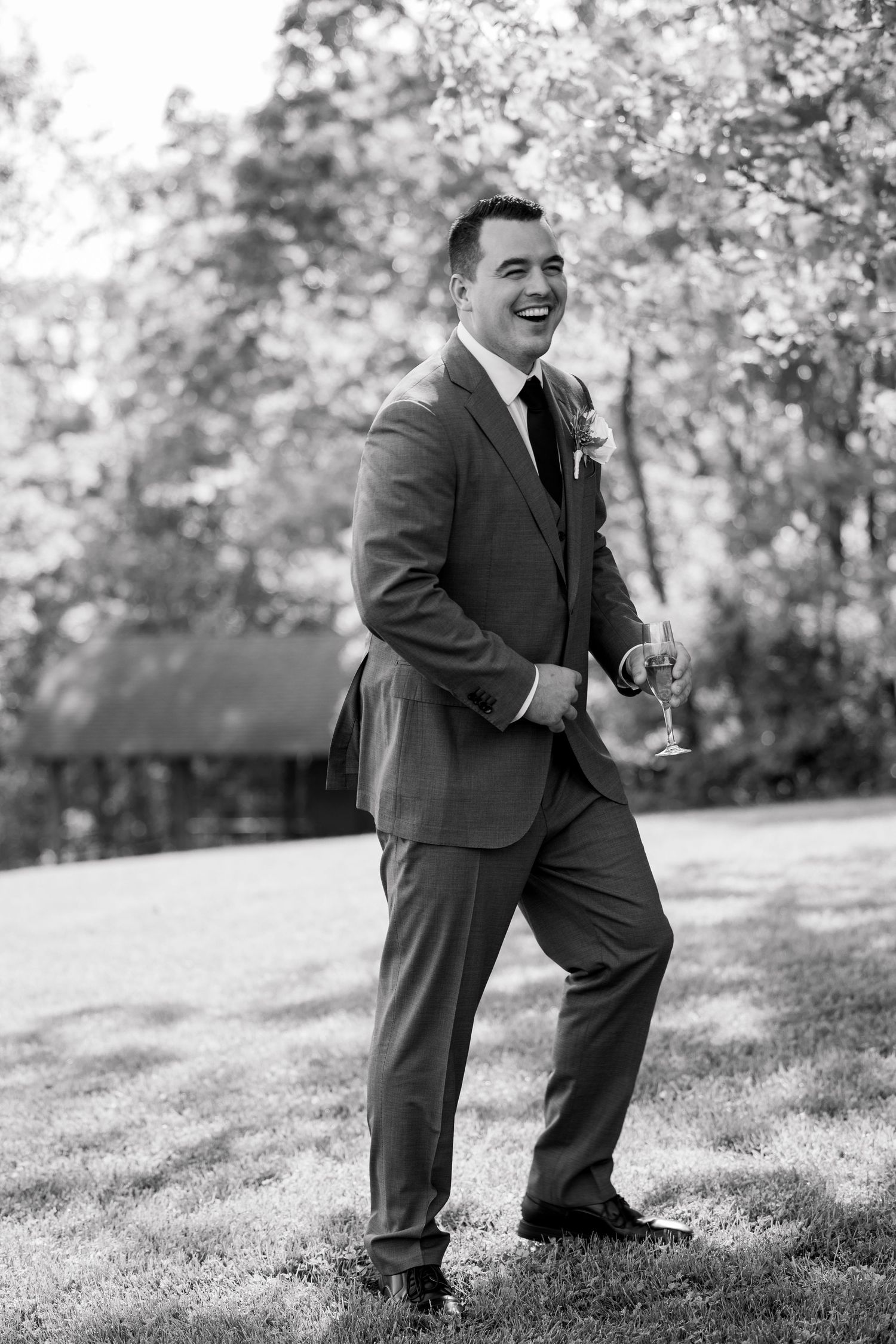 A joyful person in a gray suit smiles while standing on a grassy lawn during what appears to be a wedding day.