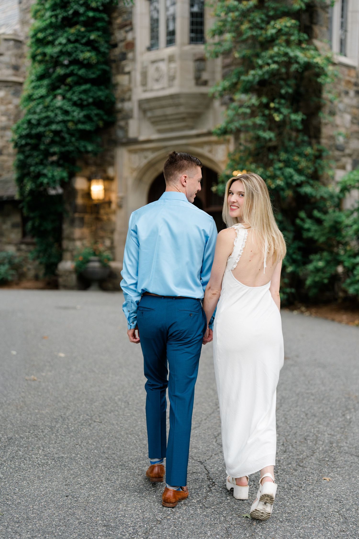 Couple walking hand in hand toward a stone castle entrance, woman in white gown and man in blue suit.