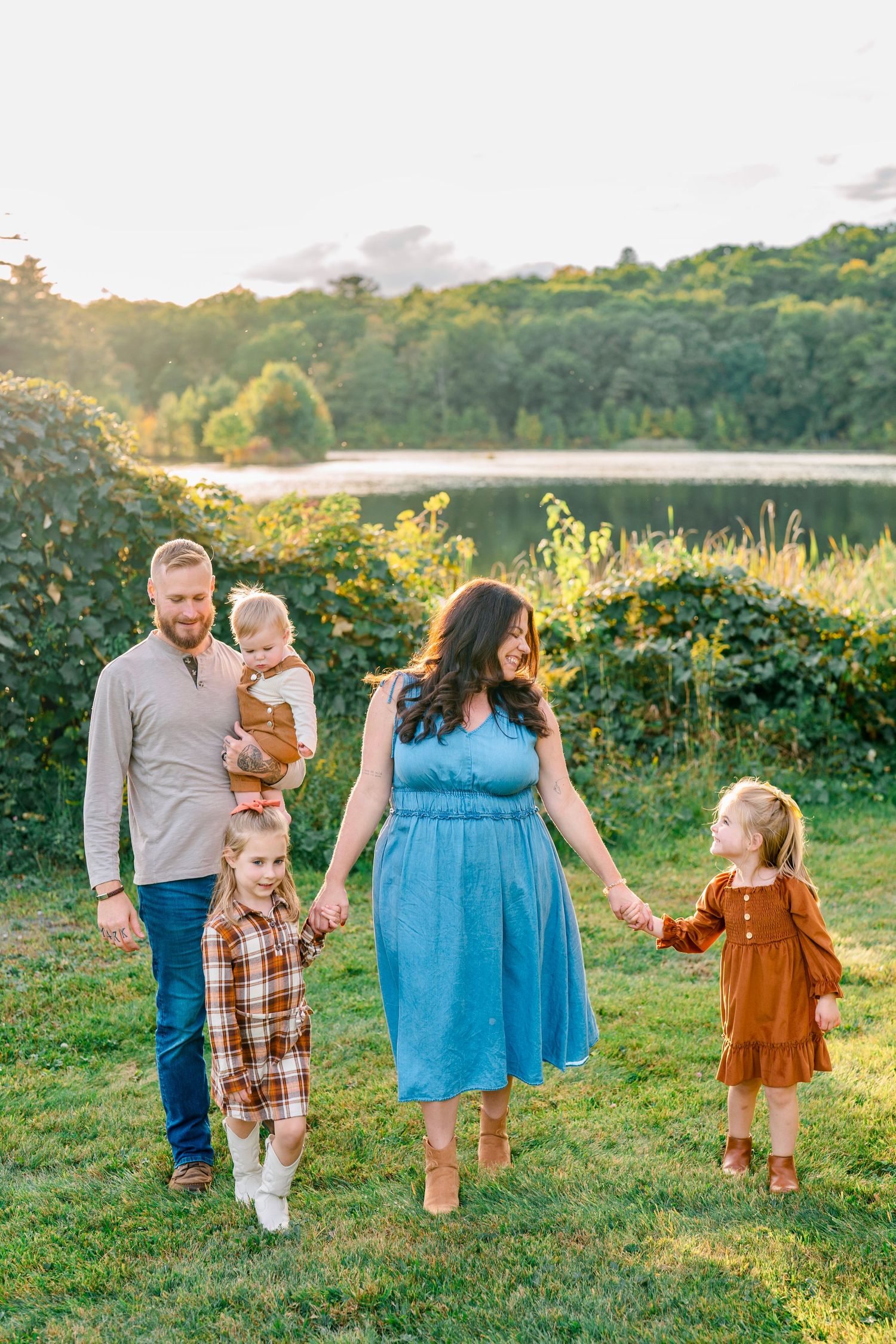 Family of five walking outdoors near a lake at golden hour, parents holding children's hands on green grass.