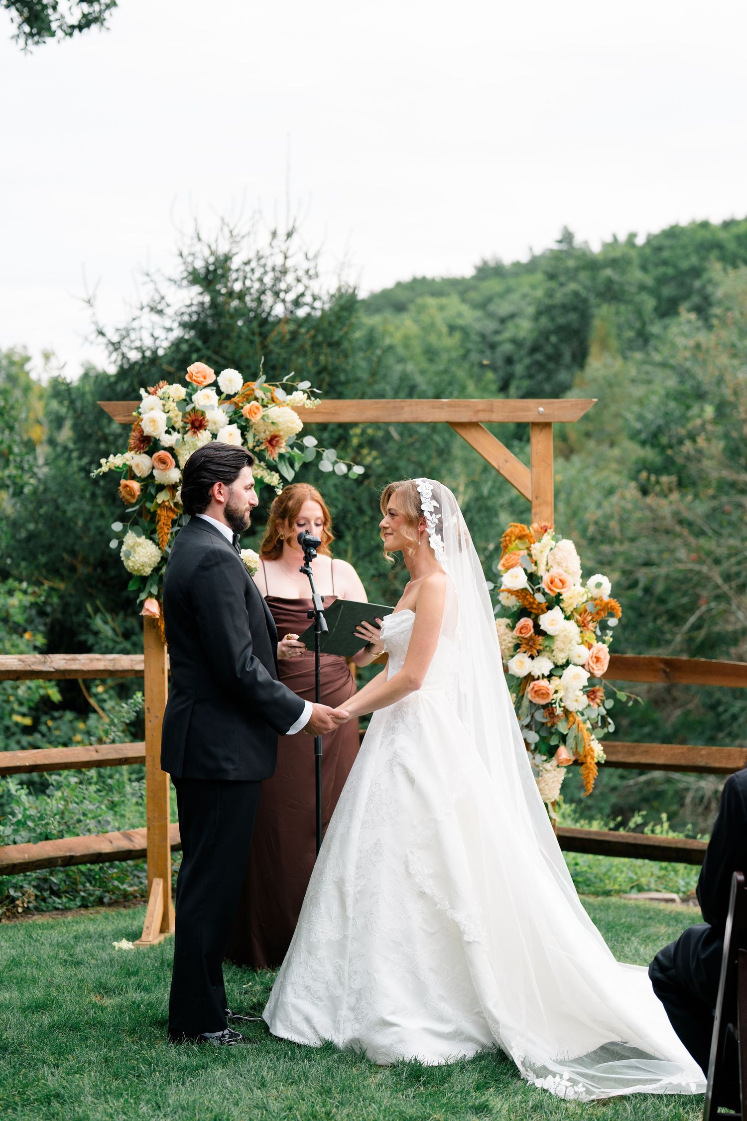 Bride and groom exchange vows under rustic wooden arch adorned with orange and white floral arrangements outdoors.