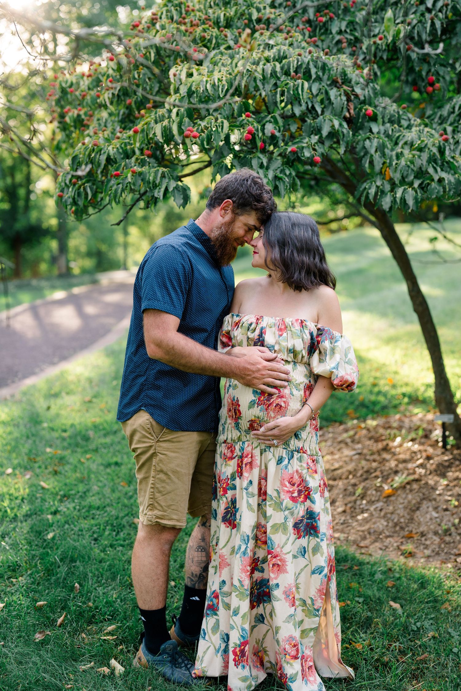 Couple tenderly embracing in a garden, man touching pregnant woman's belly in floral maxi dress under cherry tree.