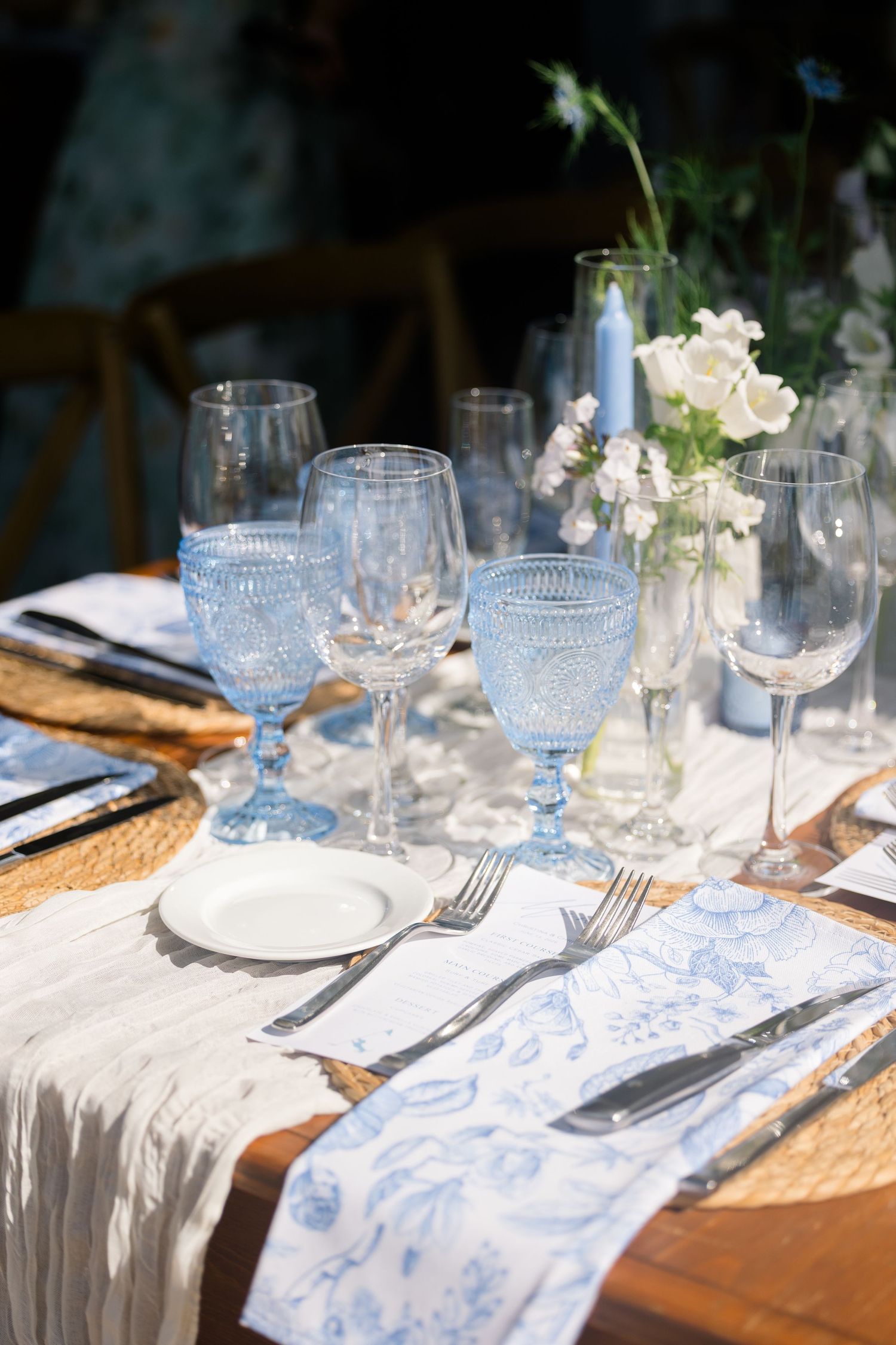 Elegant wedding table setting with blue goblets, white linens, and floral centerpieces in sunlight.