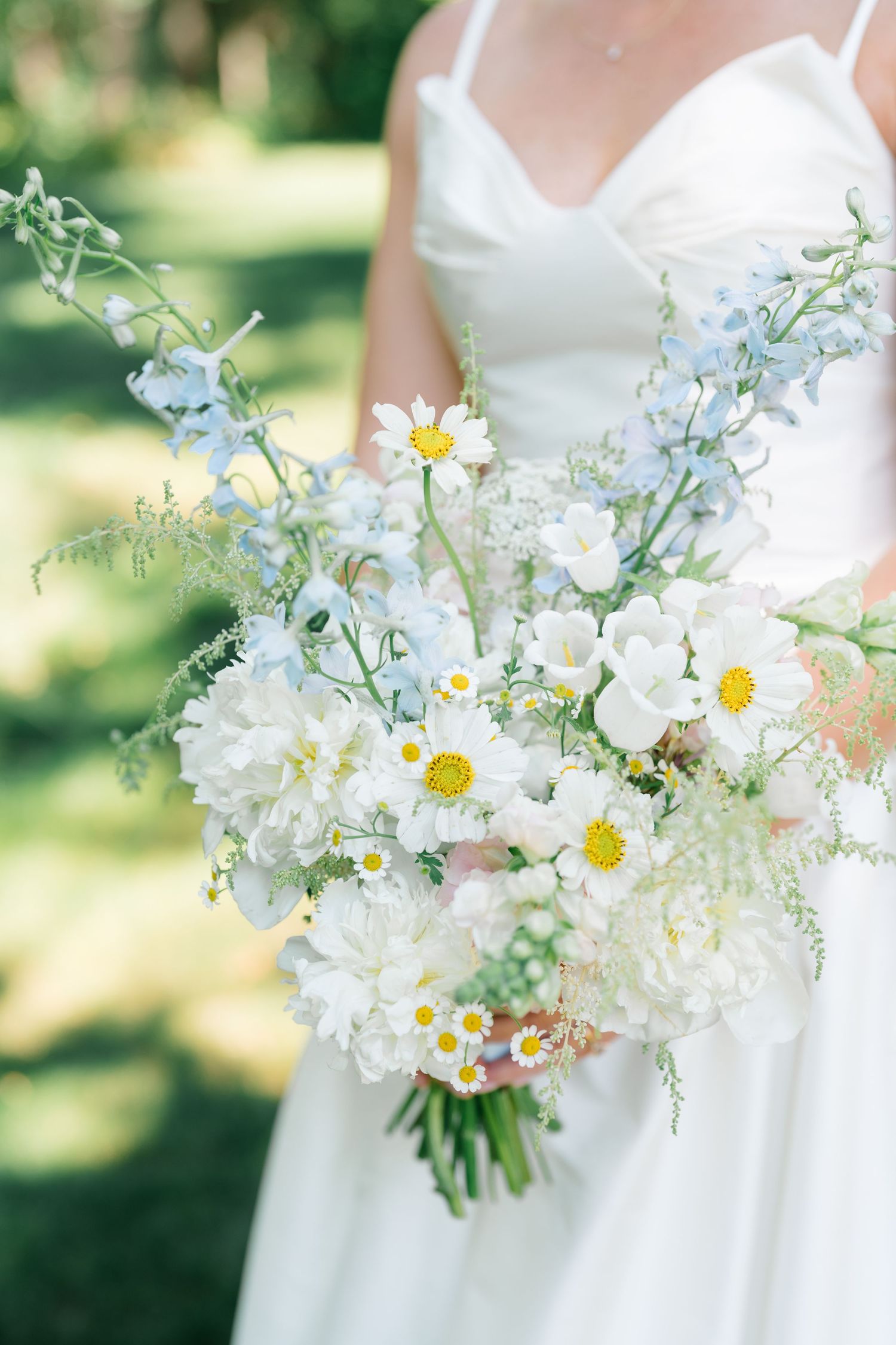 Bride in white dress holding a lush bouquet of white daisies, cosmos, hydrangeas, and blue delphinium flowers.