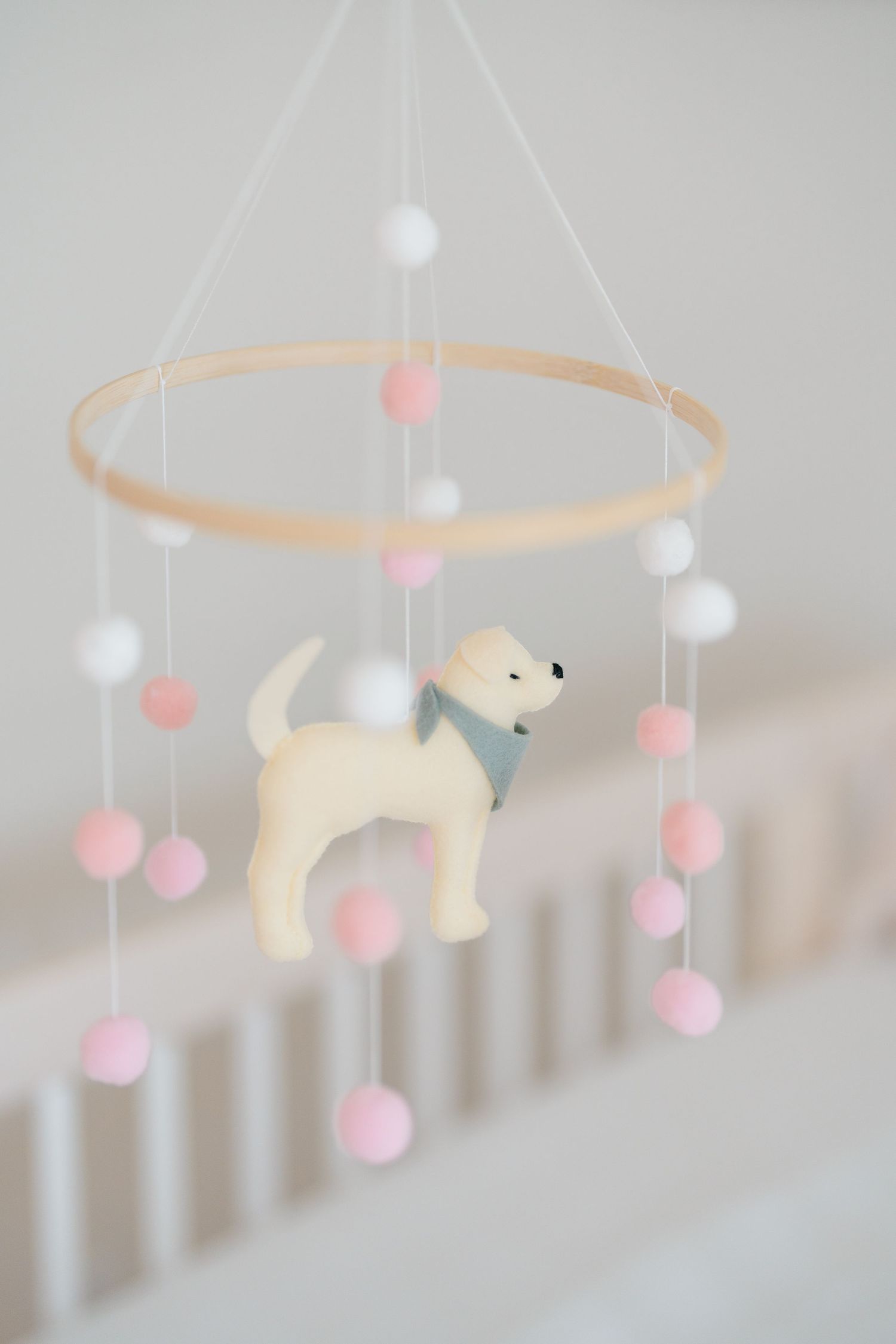 Nursery mobile featuring a white dog figurine with pink and white pom poms hanging from a wooden hoop.