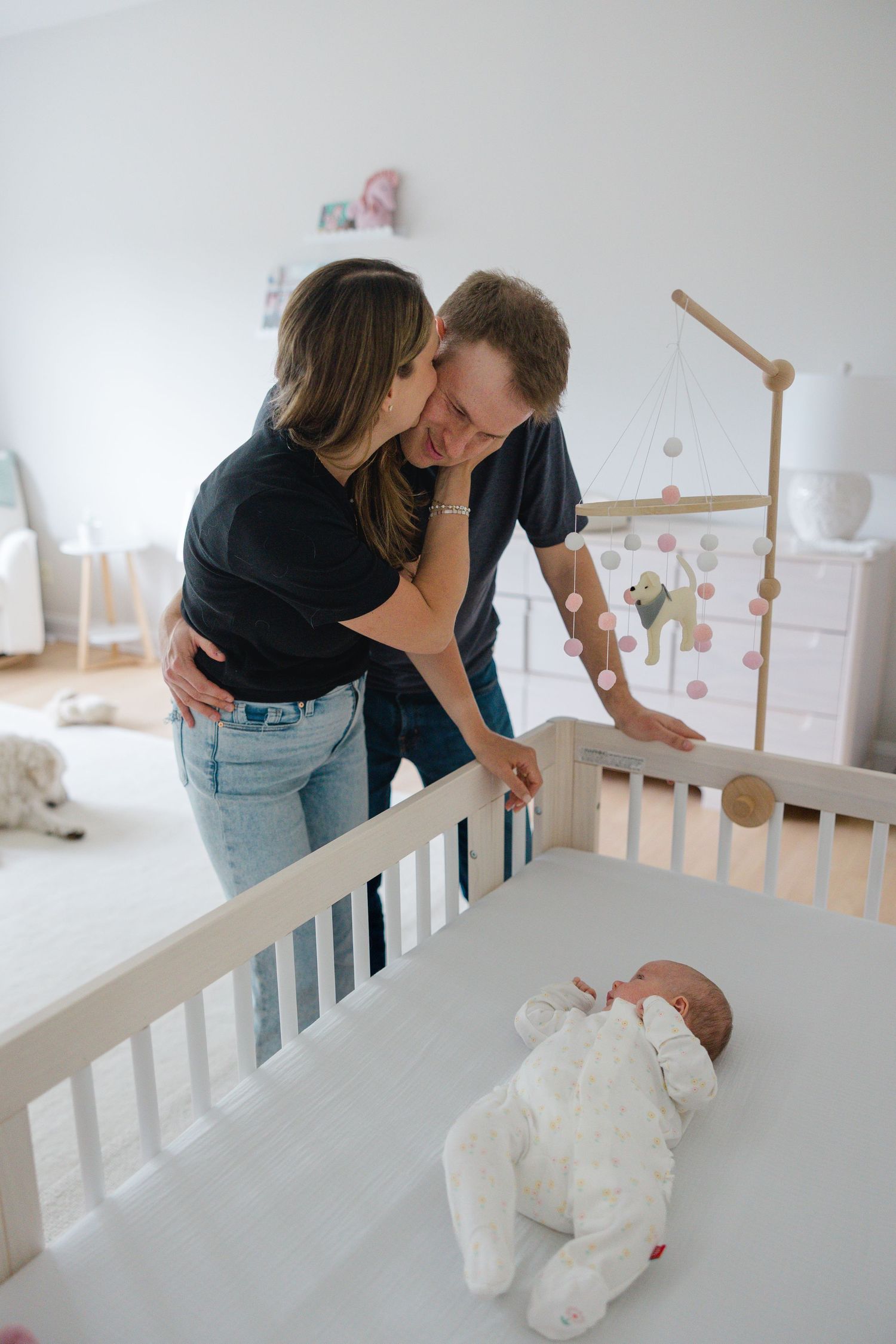 Parents lean over a white crib to share an affectionate moment while their baby rests below.