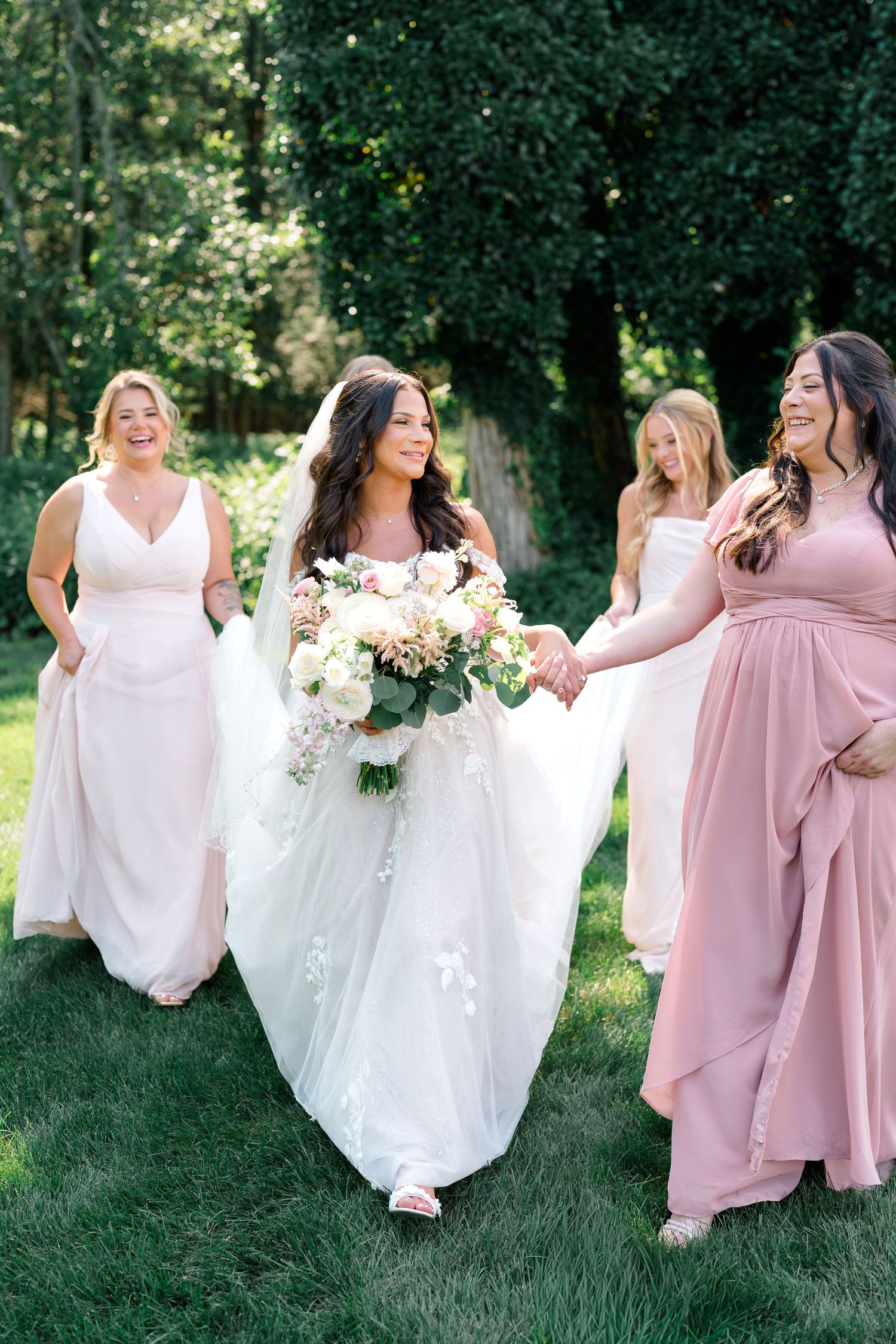 Bridesmaids in flowing pink dresses walking with bride in white gown on green lawn.
