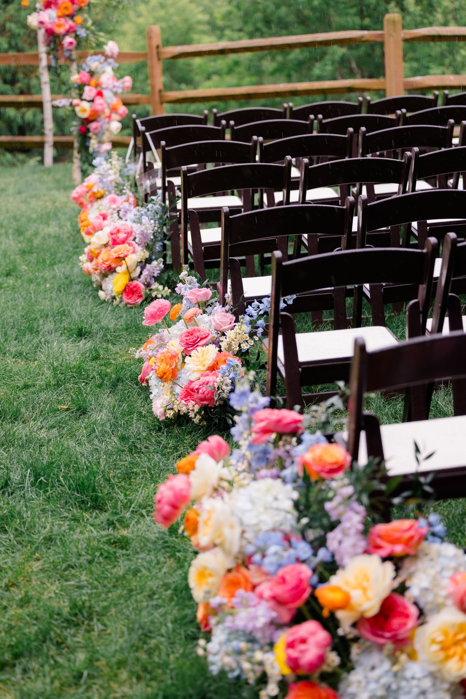 Row of dark wooden chairs lined with vibrant pink, orange and purple floral arrangements for outdoor wedding.