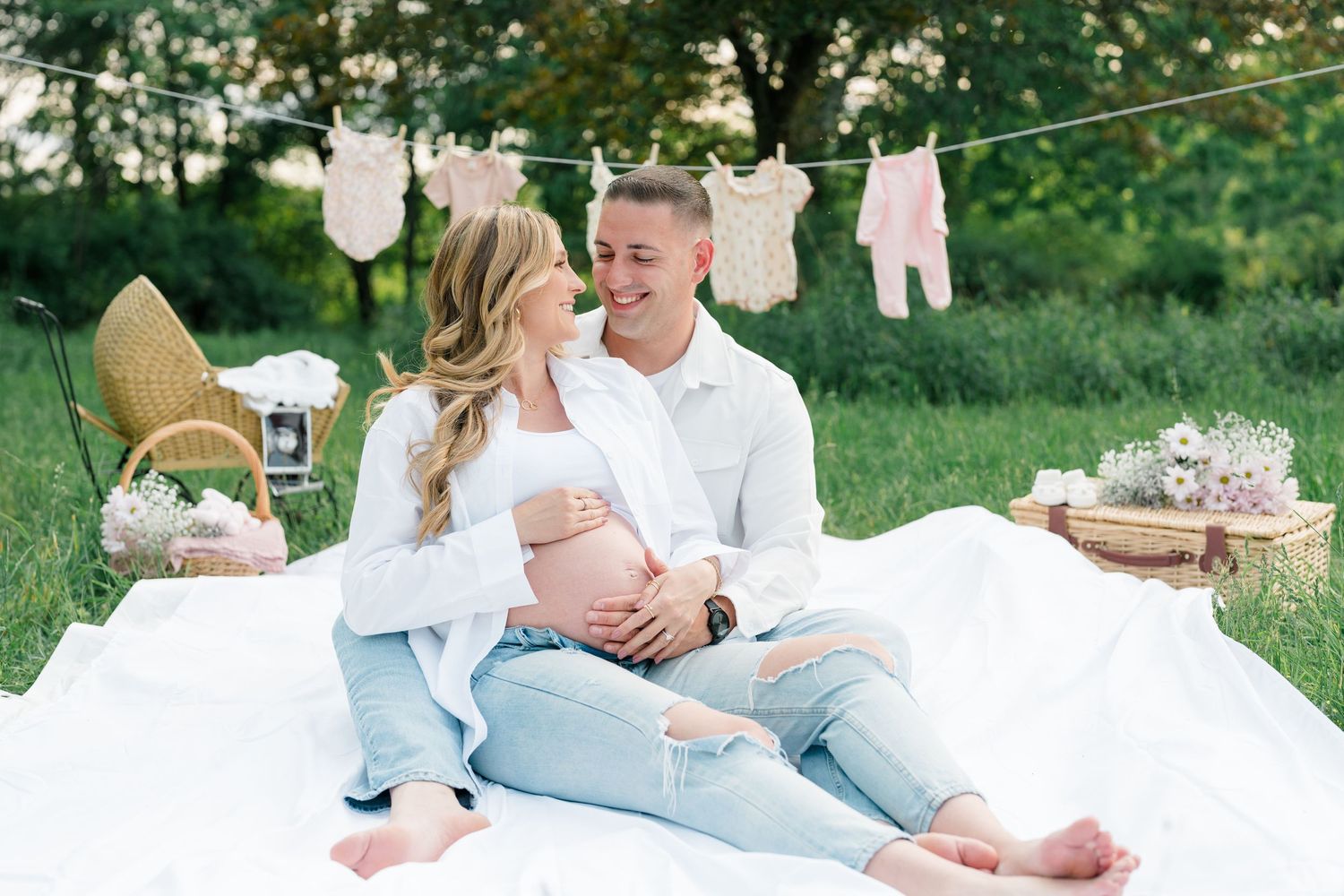 Couple lounging on white blanket outdoors with baby clothes hanging on clothesline in background.