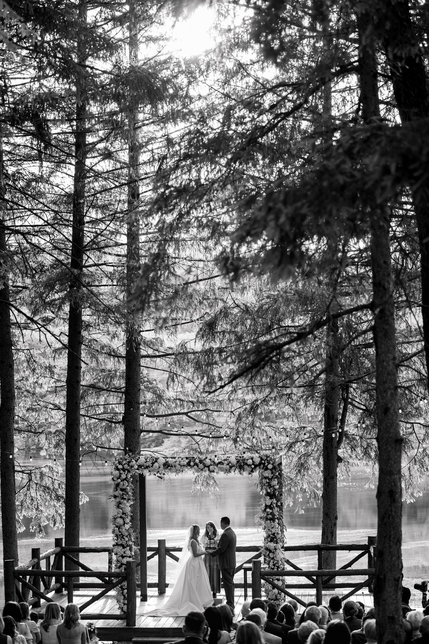 Wedding ceremony setup in a forest with wooden benches and floral arch overlooking a serene lake.